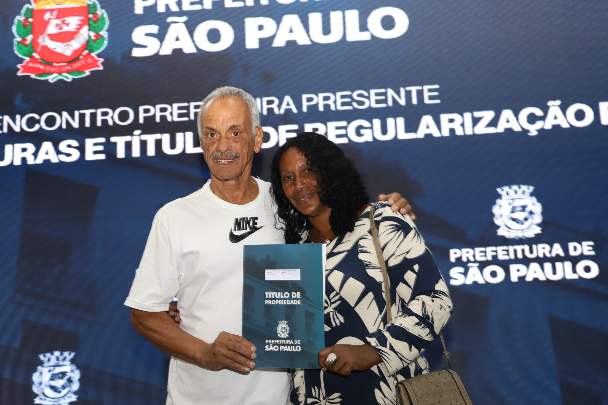 Casal sorridente segura o título de propriedade entregue pela Prefeitura de São Paulo durante evento oficial, com o painel da prefeitura ao fundo