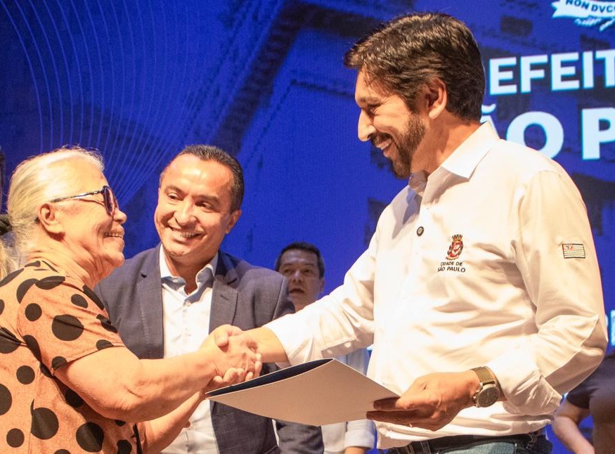 Mulher recebendo seu título de propriedade enquanto aperta a mão do prefeito Ricardo Nunes com o secretario Sidney Cruz ao fundo.