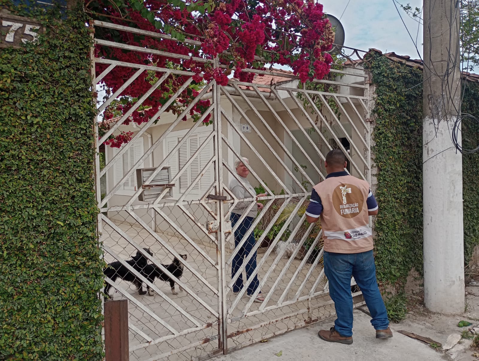 Um agente da Prefeitura de São Paulo, com colete escrito “Regularização Fundiária”, conversa com uma moradora em frente ao portão de sua casa. Um cachorro preto aparece no quintal, atrás da grade.