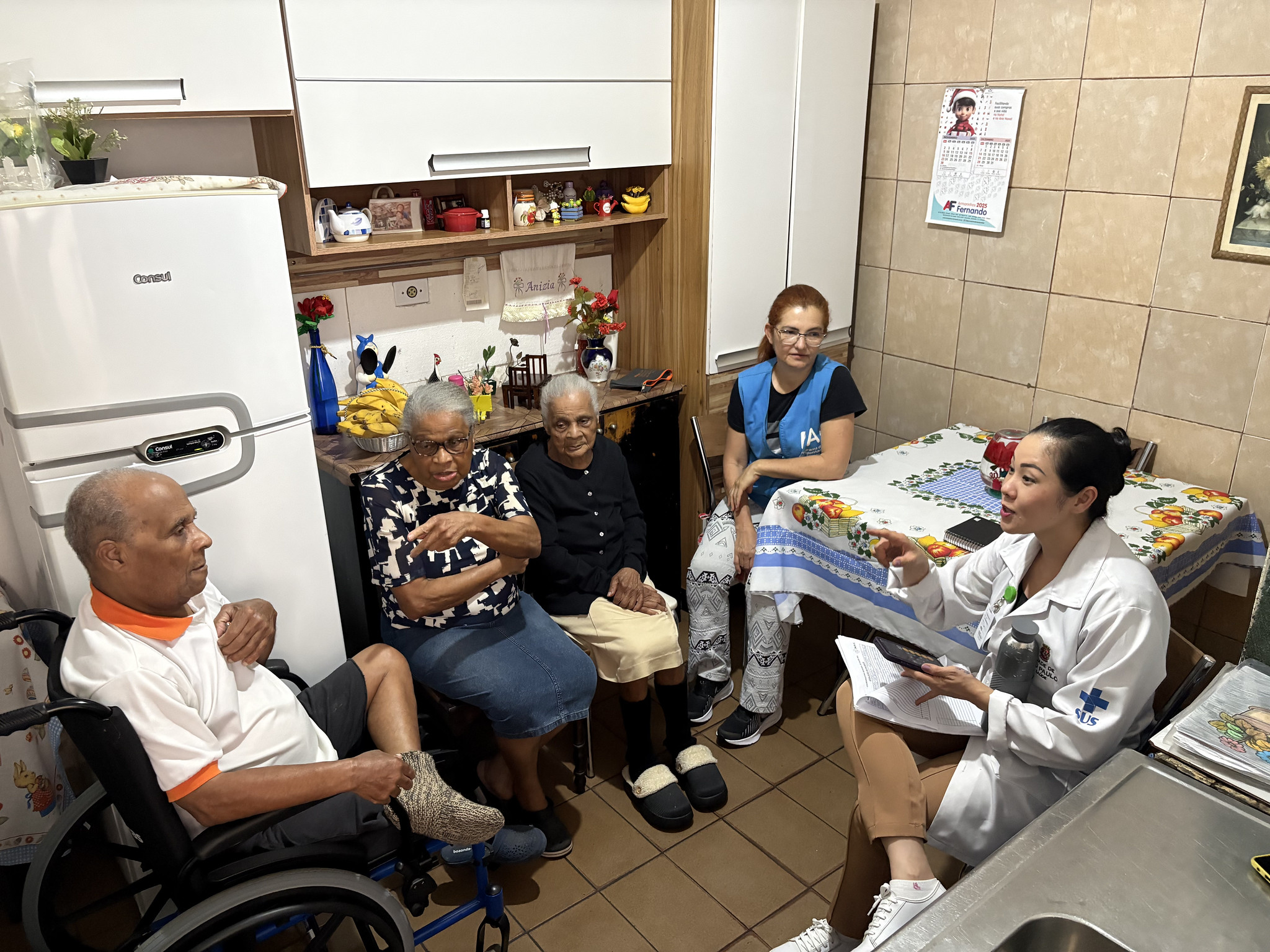A foto registra um atendimento de saúde domiciliar em uma cozinha residencial simples e iluminada. Cinco pessoas estão no ambiente: três idosos (dois sentados e um em cadeira de rodas) e duas profissionais de saúde do Programa de Acompanhante de Idosos. O grupo está organizado em um semicírculo, sugerindo uma conversa atenciosa. As Pessoas (da esquerda para a direita) Homem Idoso: Está sentado em uma cadeira de rodas manual, de perfil para a direita. Ele é negro, tem cabelos curtos grisalhos e usa uma camisa polo branca com gola laranja e bermuda escura. Ele ouve atentamente o que a profissional de saúde diz.  Mulher Idosa (ao centro): Está sentada em uma cadeira comum. Ela é negra, usa óculos de grau e uma blusa estampada em preto e branco. Ela aponta com o dedo indicador em direção à profissional, como se estivesse explicando algo.  Mulher Idosa (à direita do centro): Uma senhora negra, com cabelos brancos presos, usando uma blusa preta de manga longa, saia bege e pantufas pretas. Ela observa a cena de forma serena.  Agente de Saúde: Uma mulher de pele clara e cabelos ruivos amarrados, sentada ao fundo, entre as idosas e a mesa. Ela veste uma camiseta preta sob um colete azul (uniforme de agente de saúde) e calça estampada.  Profissional de Saúde (Médica ou Enfermeira): Sentada à direita, em primeiro plano e de perfil para a esquerda. Ela é uma mulher jovem de traços orientais, com o cabelo preso em um coque. Veste um jaleco branco com o logotipo do SUS (Sistema Único de Saúde) no braço. Ela segura um celular e papéis, gesticulando com a mão direita enquanto fala com os idosos.