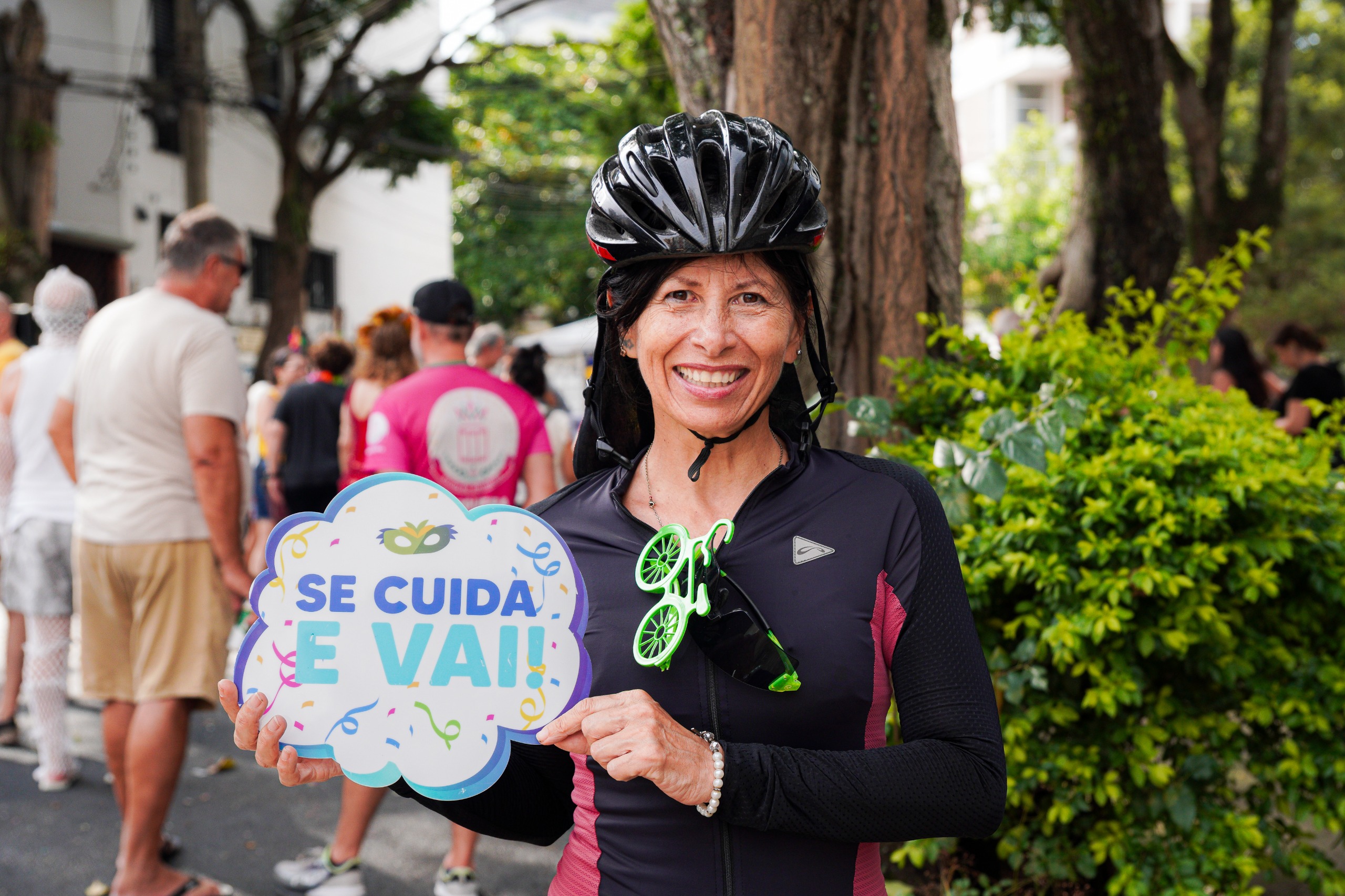 A imagem mostra uma mulher sorridente em um ambiente ao ar livre, de um bloco de Carnaval. Ela está usando capacete de ciclismo preto e roupa esportiva, indicando que está participando de uma atividade de bicicleta. No peito, há um adereço verde em formato de bicicleta preso à roupa. Ela segura uma placa decorativa em formato de nuvem com a frase “SE CUIDA E VAI!” escrita em letras coloridas, acompanhada de pequenos desenhos festivos. Ao fundo, há várias pessoas caminhando, além de árvores grandes, arbustos verdes e prédios residenciais.