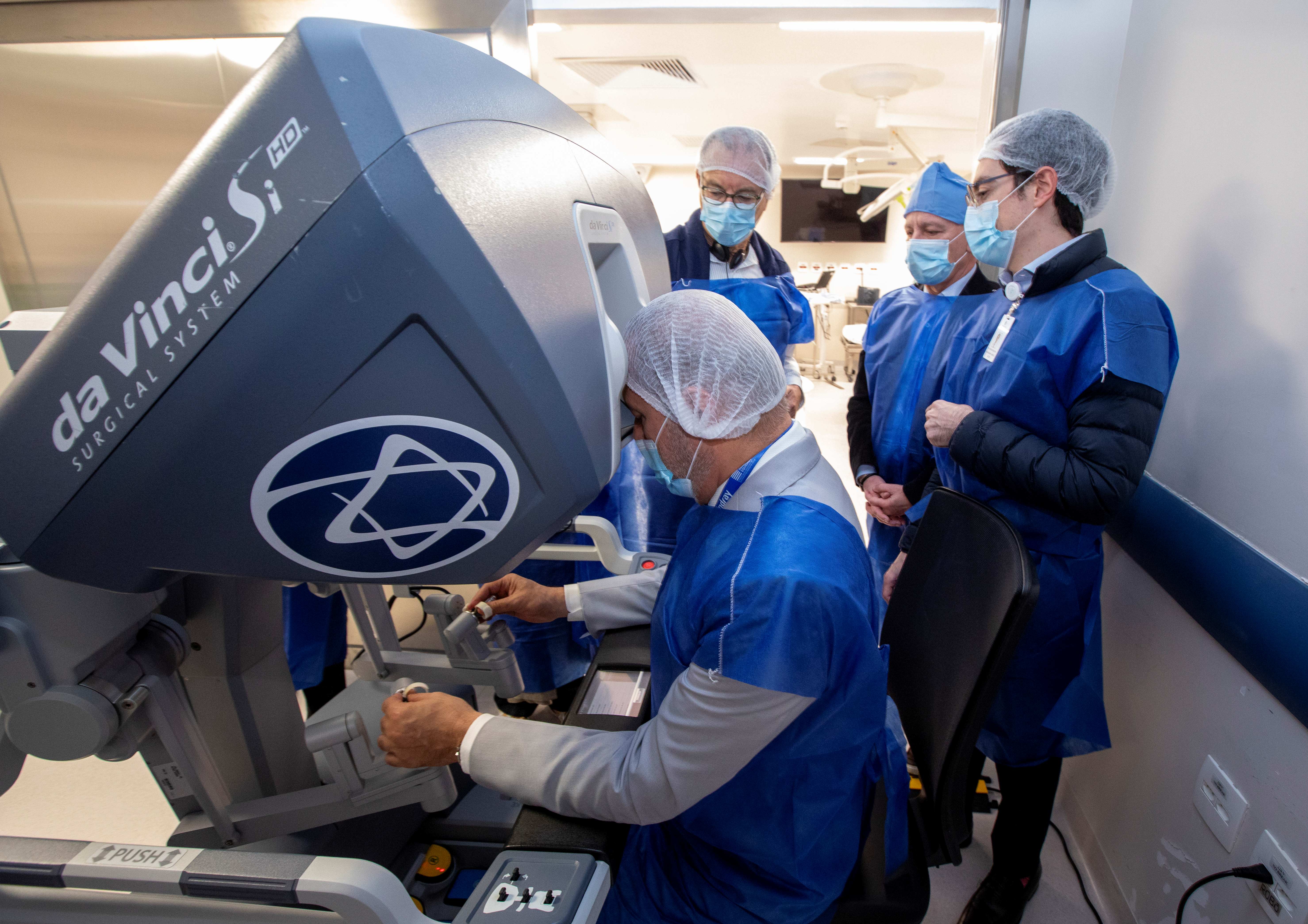 A imagem mostra uma cirurgia robótica em um ambiente hospitalar ou de centro cirúrgico. Quatro pessoas estão utilizando roupas e equipamentos de proteção: aventais azuis descartáveis, toucas e máscaras.  No primeiro plano, uma pessoa está sentada e operando um equipamento grande e moderno, que é um sistema de cirurgia robótica. Essa pessoa olha para dentro do visor da máquina e manuseia dois controles com as mãos, como se estivesse treinando ou realizando um procedimento por meio do robô. Ao redor dela, outras três pessoas observam atentamente o processo. 