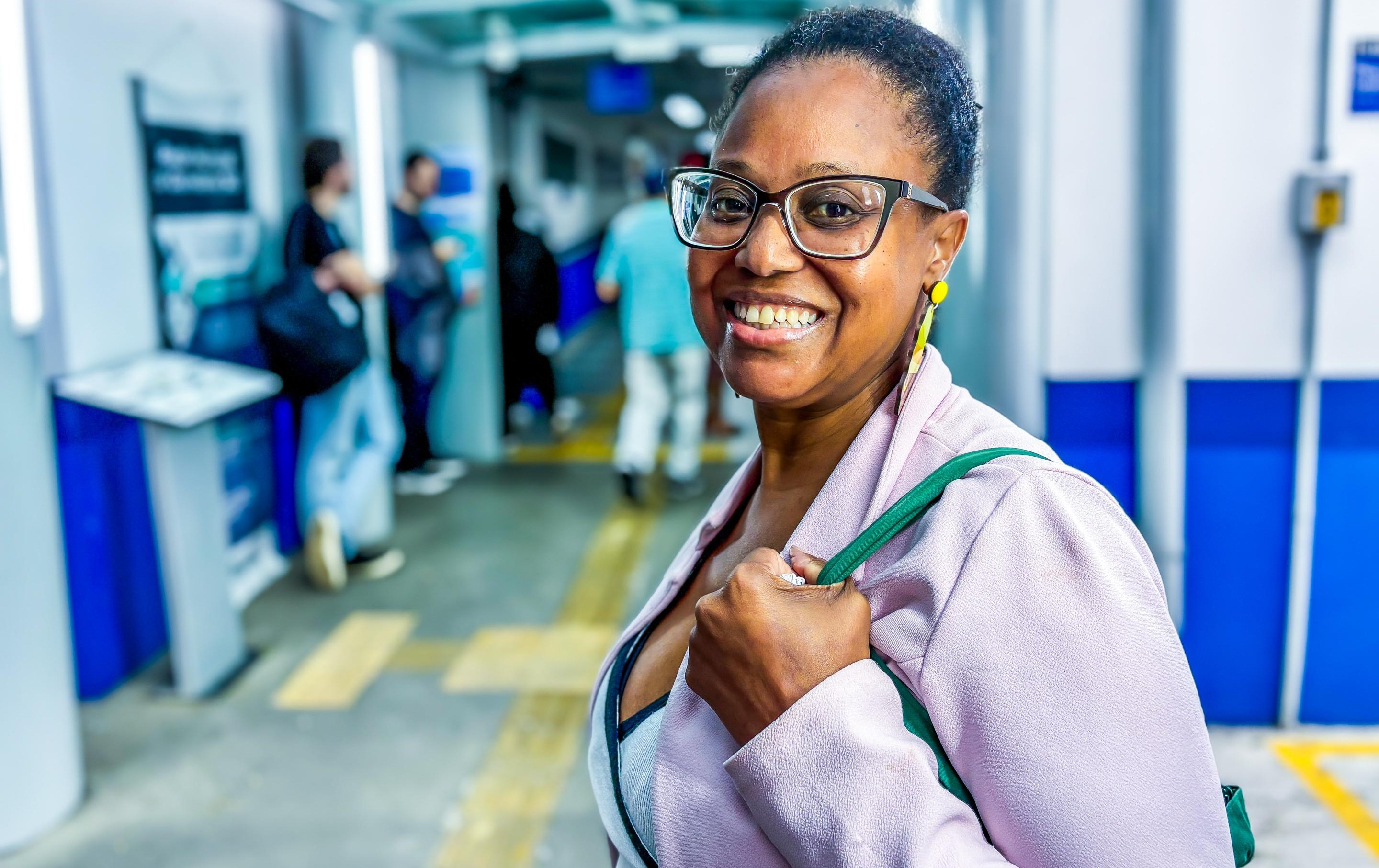 A imagem mostra uma mulher sorridente, de pele negra, usando óculos de armação grossa e brincos amarelos. Ela está vestida com um casaco claro e carrega uma bolsa verde sobre o ombro. O fundo está desfocado, mas é possível ver que ela está em um ambiente interno, com paredes brancas e azuis, e algumas pessoas ao fundo, sugerindo um espaço de circulação pública, como uma unidade de saúde ou um corredor de hospital. A iluminação é clara, e a expressão da mulher transmite simpatia e confiança