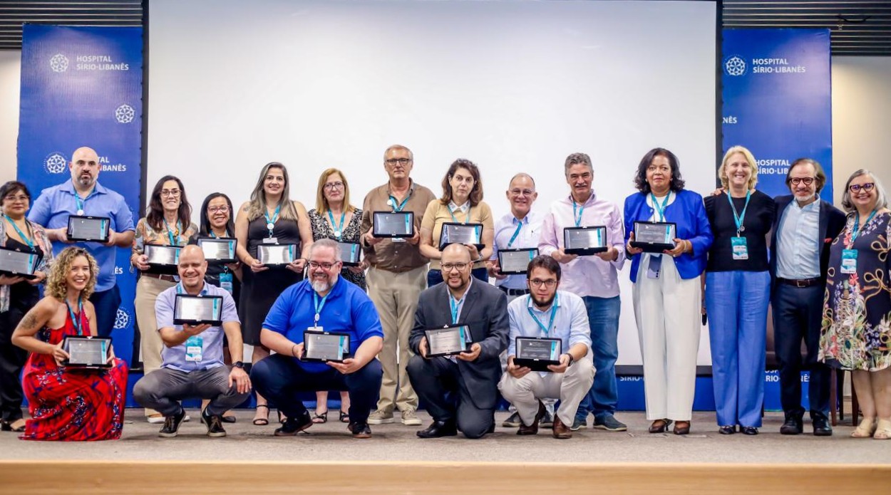 A imagem mostra um grupo de pessoas posando em um auditório, em frente a um painel com o logotipo do Hospital Sírio-Libanês. Elas estão sorridentes e seguram placas de premiação ou certificados, indicando uma cerimônia de reconhecimento ou homenagem.  Há cerca de vinte pessoas — parte delas em pé e parte agachada na frente —, todas usando crachás com cordões azul-turquesa. O ambiente é bem iluminado, com fundo branco e banners azuis laterais com o nome e o símbolo do hospital. A atmosfera é de celebração e conquista profissional.