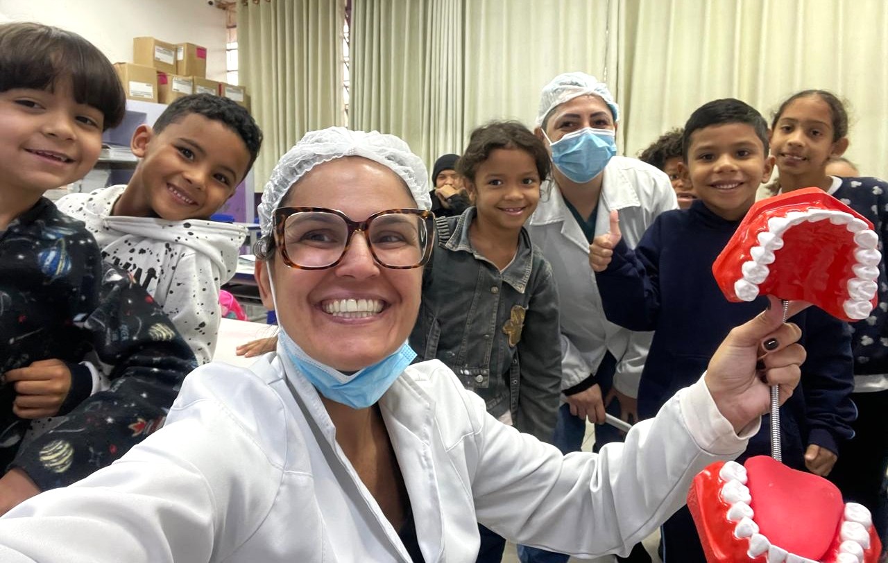 A imagem mostra um grupo sorridente composto por duas profissionais da área da saúde e várias crianças. Uma das profissionais, que usa jaleco branco, touca e óculos, segura um grande modelo didático de boca com dentes, utilizado para demonstrar técnicas de higiene bucal. As crianças, que aparentam estar em idade escolar, posam animadas ao redor das profissionais. O ambiente parece ser uma sala de aula ou um espaço educativo, com cortinas claras e caixas empilhadas ao fundo. A cena transmite alegria e interação, sugerindo uma atividade de educação em saúde bucal.
