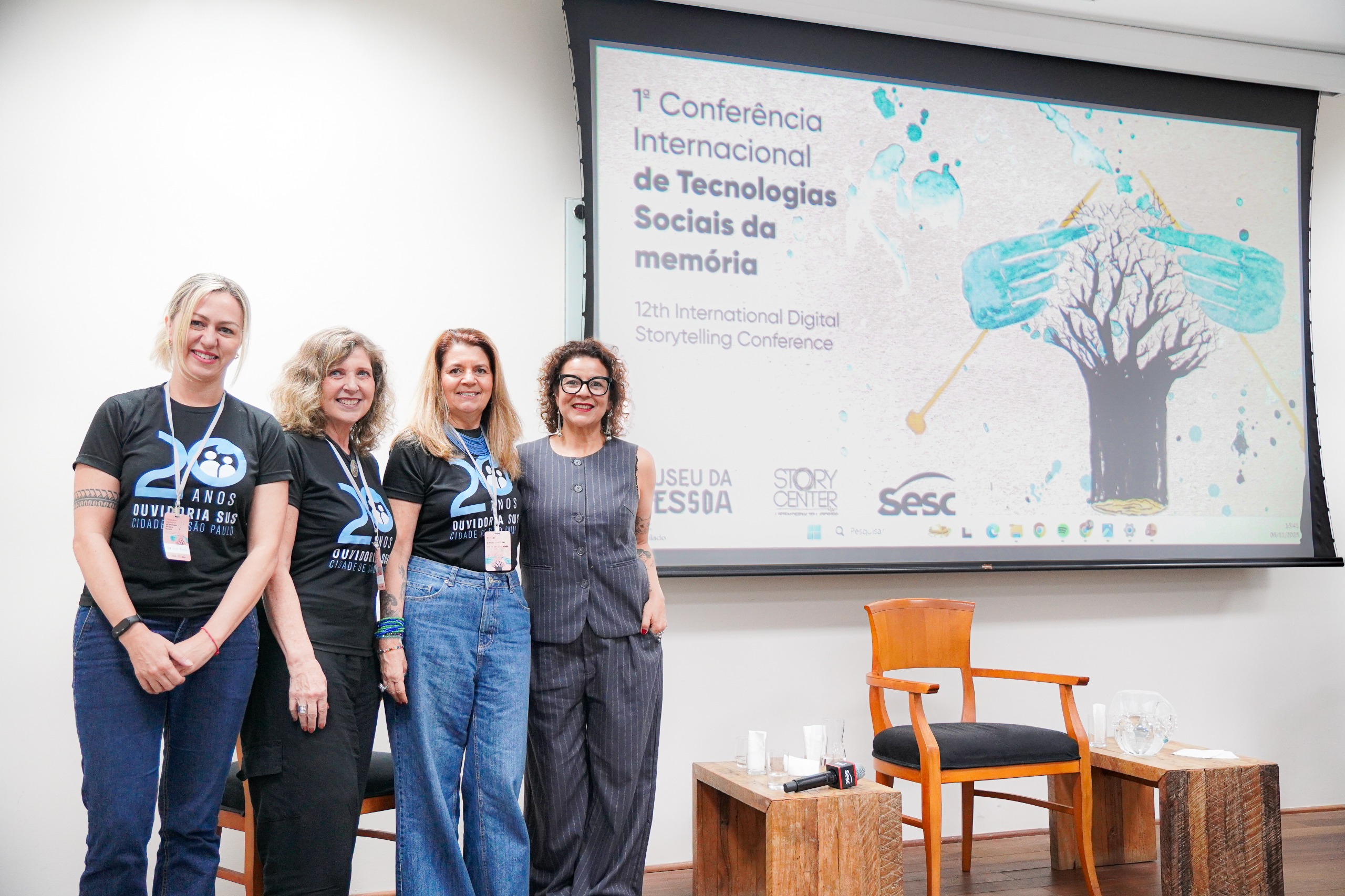 A imagem mostra quatro mulheres posando sorridentes diante de uma tela de projeção em um auditório. Três delas vestem camisetas pretas com o logotipo dos 20 anos da Ouvidoria SUS – Cidade de São Paulo, e a quarta usa um conjunto social cinza. Atrás delas, o slide projetado exibe o título “1ª Conferência Internacional de Tecnologias Sociais da Memória – 12th International Digital Storytelling Conference”, acompanhado dos logotipos do Museu da Pessoa, StoryCenter e Sesc. À frente, há duas cadeiras de madeira e uma pequena mesa com microfone, copos e uma jarra de água, compondo o ambiente de uma apresentação ou painel do evento.