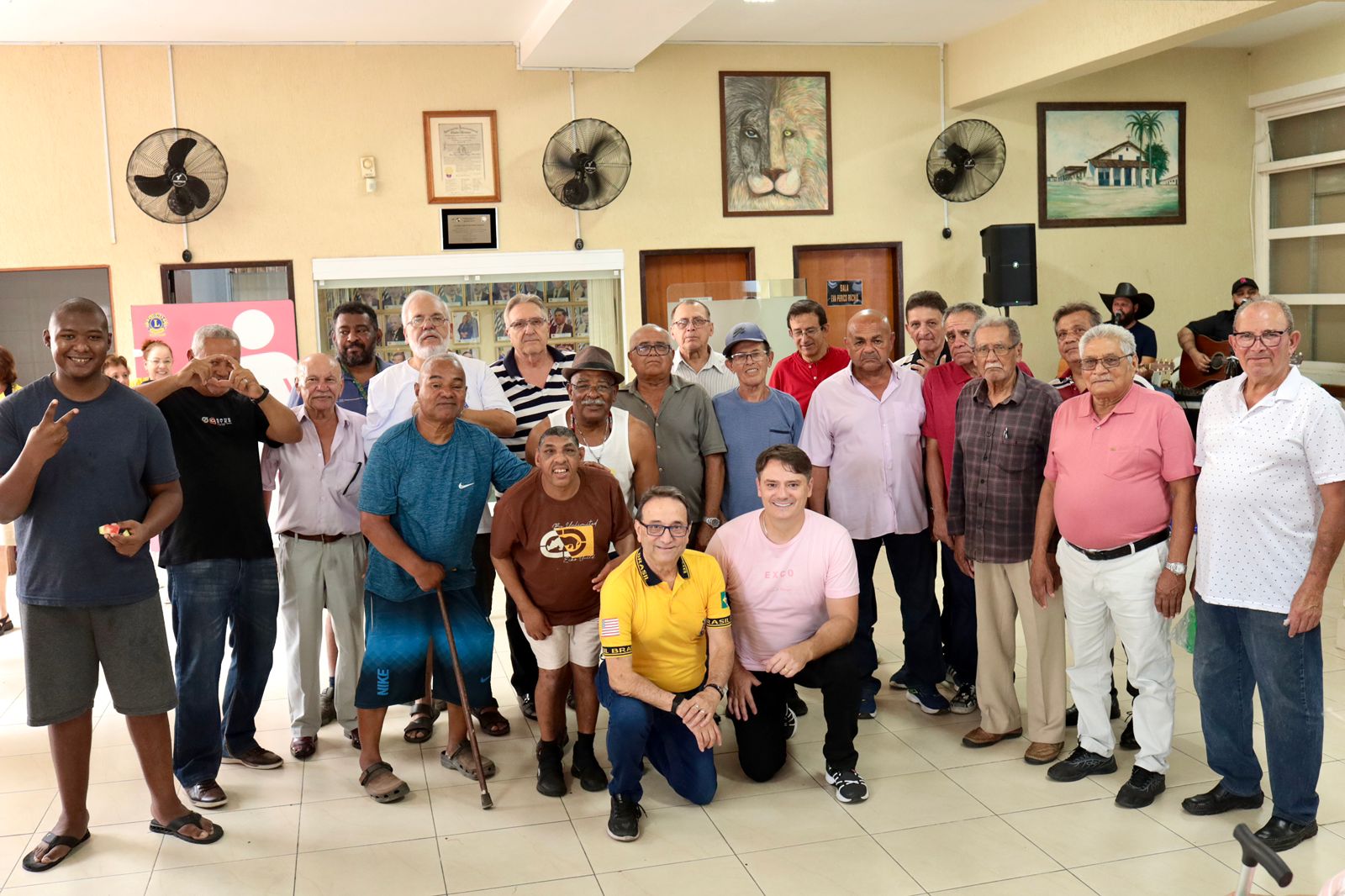 A imagem mostra um grupo grande de homens adultos, a maioria de meia-idade ou mais velhos, posando para uma foto. Há cerca de 23 homens visíveis. O vestuário é casual, variando de camisetas e camisas polo a camisas de botões. Muitos estão sorrindo ou olhando para a câmera. Um homem no canto esquerdo está fazendo um sinal de 