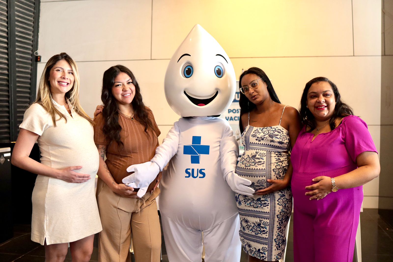 A imagem mostra quatro mulheres grávidas posando para a foto ao lado do mascote do SUS, o Zé Gotinha.  Todas estão sorrindo ou demonstrando expressão amistosa, com as mãos apoiadas nas próprias barrigas, em um gesto típico de gestantes. O Zé Gotinha está no centro, com sua roupa branca característica, símbolo azul do SUS no peito e um grande sorriso no rosto.  As mulheres vestem roupas confortáveis em tons claros e vibrantes — bege, marrom, azul estampado e rosa. O ambiente parece ser interno, bem iluminado, provavelmente relacionado a uma ação de saúde ou vacinação, já que há um cartaz do SUS ao fundo. 