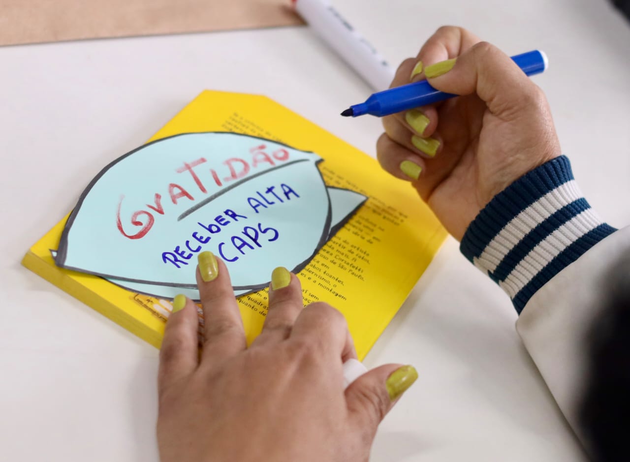 A imagem mostra um close das mãos de uma pessoa sobre uma mesa branca. Ela segura uma caneta azul e escreve em um recorte de papel colorido, em formato arredondado, colocado sobre um livro amarelo. No papel está escrito à mão 
