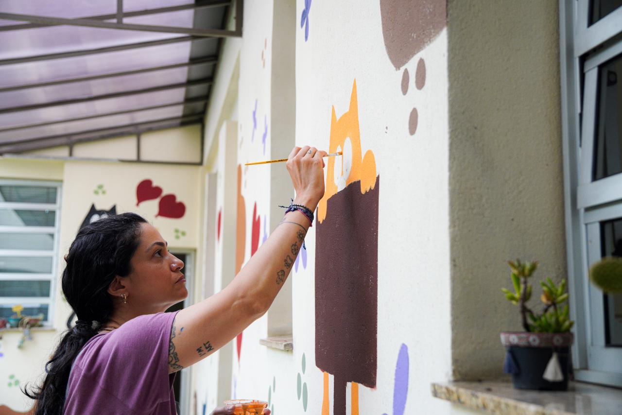 A imagem mostra uma mulher pintando um mural em uma parede externa, possivelmente em um ambiente escolar ou comunitário. Ela segura um pincel com a mão direita e aplica tinta em um desenho colorido, que parece representar personagens ou formas lúdicas. Na outra mão, segura um pequeno recipiente com tinta.  A mulher veste uma camiseta roxa, tem tatuagens nos braços e o cabelo preso. Sua expressão é concentrada, indicando atenção aos detalhes do trabalho. Ao fundo, há janelas, vasos de plantas e outras pinturas na parede, com corações e figuras decorativas, reforçando o caráter artístico e acolhedor do espaço.