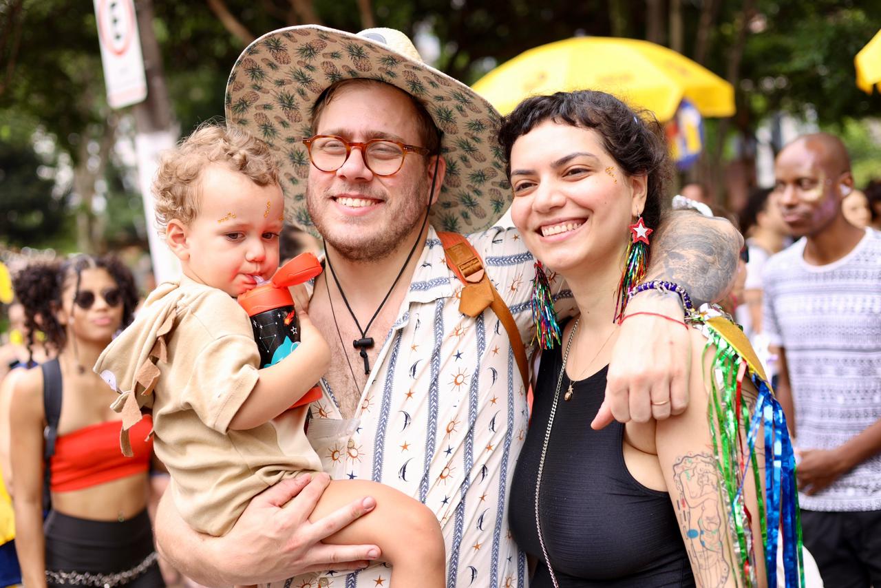 A imagem mostra três pessoas em primeiro plano, em meio a um ambiente festivo ao ar livre, que parece ser um bloco de Carnaval ou uma celebração popular.  No centro, há um homem sorrindo, usando óculos, chapéu estampado e camisa clara com desenhos. Ele segura no colo uma criança pequena, que parece estar bebendo água de uma garrafinha vermelha. A criança veste uma roupa bege e tem glitter no rosto, típico de festas carnavalescas.  À direita, há uma mulher sorridente, com o braço em volta do homem. Ela usa uma blusa preta sem mangas, brincos coloridos em forma de estrela e tem tatuagens visíveis no braço. Também carrega enfeites coloridos pendurados no ombro, reforçando o clima festivo.  Ao fundo, aparecem outras pessoas participando do evento, além de árvores e guarda-sóis amarelos,