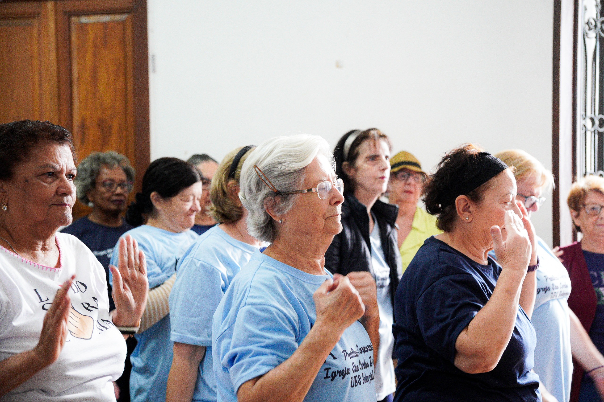 A cena retrata usuárias da UBS Belenzinho participando das atividades físicas da Academia da Saúde, iniciativa que promove práticas corporais, convivência e bem-estar. A atividade simboliza o fortalecimento das ações de promoção da saúde na cidade, em um momento em que o programa foi ampliado, passando de 18 para 66 polos na capital paulista. Elas estão lado a lado, realizando movimentos coordenados com os braços, aparentemente seguindo orientações de uma prática de exercícios ou alongamento. Muitas usam camisetas claras — algumas em azul — possivelmente relacionadas à atividade ou ao grupo de saúde. As expressões são de concentração e engajamento, indicando atenção aos movimentos. Ao fundo, outras participantes acompanham o exercício, formando um grupo numeroso e diverso.