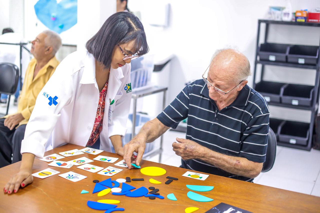 A fotografia em plano médio mostra uma interação entre uma profissional de saúde e um paciente idoso em uma sala iluminada. À esquerda, uma mulher jovem de pele parda e cabelos pretos curtos, vestindo um jaleco branco com o logotipo do SUS (Sistema Único de Saúde) no braço, está inclinada sobre uma mesa de madeira. Ela observa atentamente a atividade. À direita, um senhor idoso, calvo e de óculos, veste uma camisa polo listrada em tons de azul escuro. Ele está sentado e utiliza as mãos para organizar peças geométricas coloridas (azuis, amarelas e pretas) e cartões com ilustrações sobre a mesa. O ambiente ao fundo é clínico, com paredes brancas, uma estante de metal com gaveteiros pretos e outro paciente sentado ao longe. O foco da imagem transmite paciência, reabilitação e concentração.
