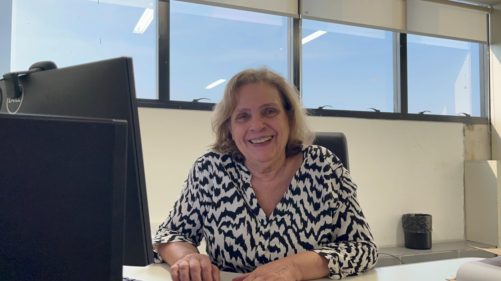 A imagem mostra a médica Flávia Terzian, uma mulher loira e de meia-idade, sentada em sua mesa de escritório e sorrindo para a câmera. Ela está usando uma blusa preta e branca com estampa abstrata. Atrás dela, há uma janela grande que dá para a rua e um computador. A imagem é bem iluminada e tem um clima descontraído e amigável.