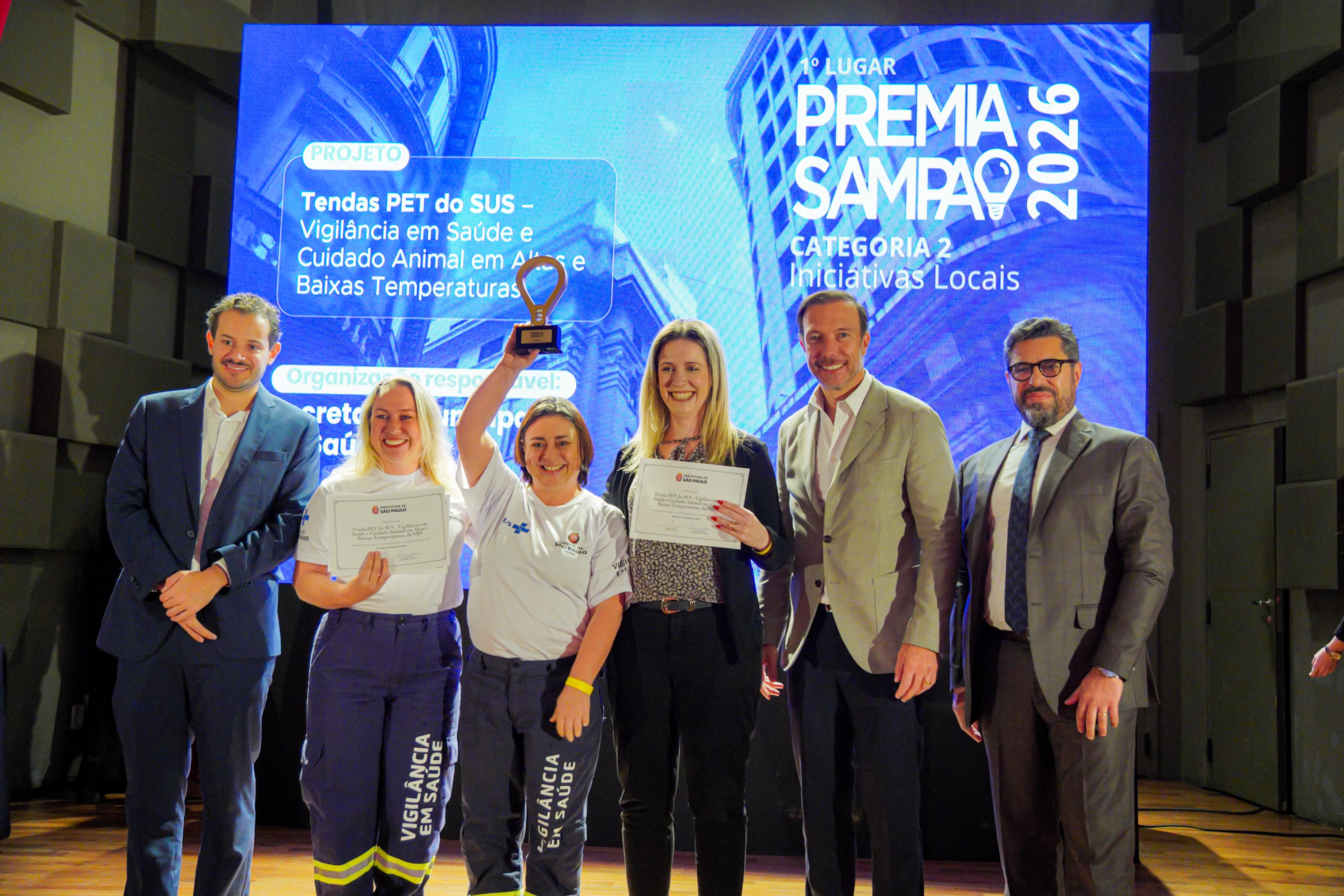 A imagem mostra uma cerimônia de premiação realizada em um auditório, com seis pessoas posando no palco diante de um telão que exibe o resultado do Premia Sampa 2026. Ao centro, integrantes da equipe vencedora seguram certificados e um troféu erguido acima da cabeça, celebrando a conquista. No telão ao fundo, é possível ler que o projeto premiado foi “Tendas PET do SUS – Vigilância em Saúde e Cuidado Animal em Ações de Baixas Temperaturas”, vencedor do 1º lugar na Categoria 2 – Iniciativas Locais. A tela também indica a vinculação do projeto à Secretaria Municipal da Saúde de São Paulo. As duas profissionais ao centro vestem camisetas da Vigilância em Saúde e exibem os certificados da premiação, enquanto os demais participantes, trajando roupas sociais, acompanham o momento de reconhecimento institucional. A iluminação do palco e a projeção ao fundo reforçam o caráter formal do evento.