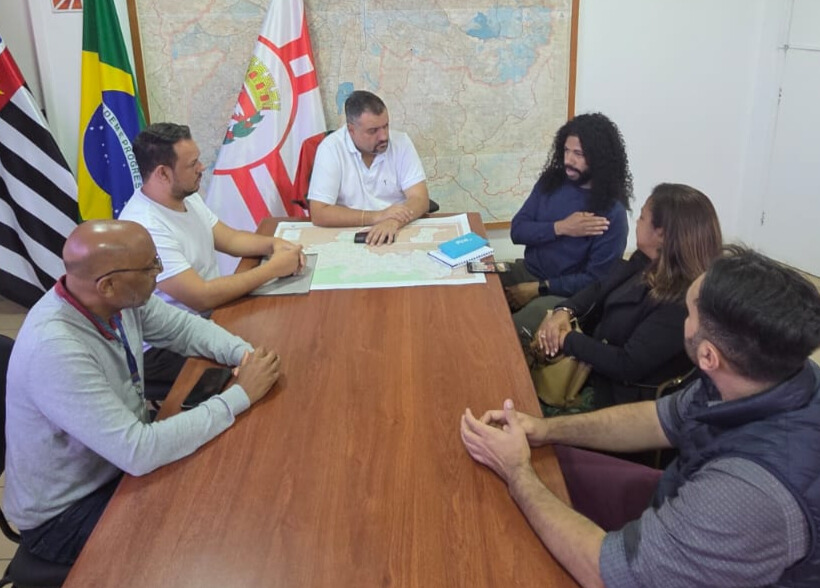 Seis pessoas estão sentadas ao redor de uma mesa retangular de madeira em uma sala de reuniões. A mesa tem sobre ela alguns cadernos, mapas e celulares. Ao fundo, há três bandeiras: do estado de São Paulo, do Brasil e da cidade de São Paulo. Também há um grande mapa na parede. No centro da mesa, um homem de camiseta branca, o subprefeito, , está com as mãos sobre o mapa. À sua direita, um homem de cabelo comprido e cacheado, usando uma blusa azul, fala .Ao lado dele, uma mulher de cabelos lisos e castanhos escuros o observa com atenção. Do outro lado da mesa, três homens — um de camiseta branca, um calvo de óculos e outro com colete escuro — acompanham a conversa. O clima sugere uma reunião institucional .