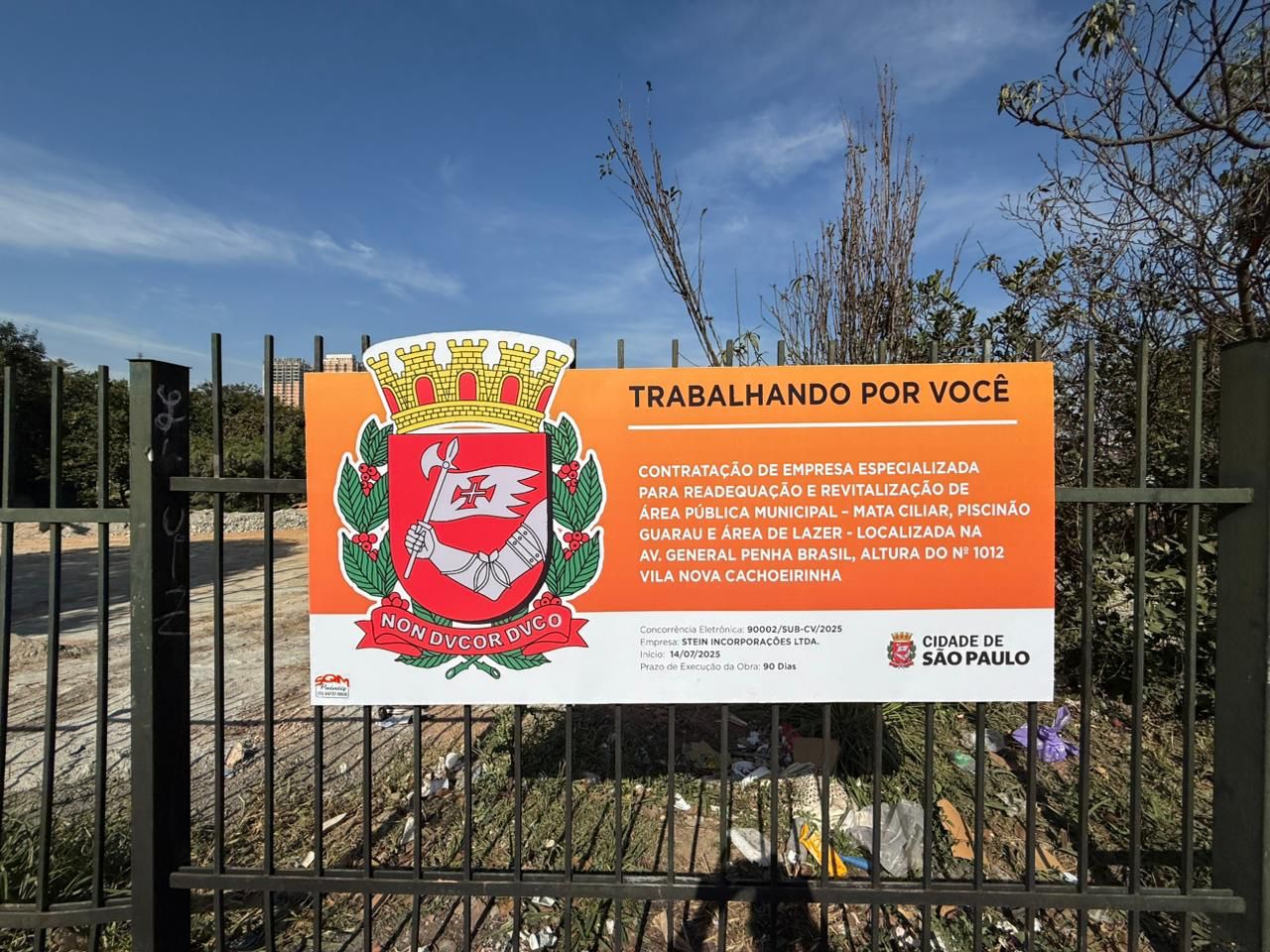 A imagem mostra uma placa afixada em uma grade de metal verde, em frente a um terreno em obras. A placa é retangular, com fundo em tons de laranja e branco, e contém informações sobre uma obra pública da Prefeitura de São Paulo. No lado esquerdo da placa está o brasão oficial da cidade de São Paulo, com um escudo vermelho contendo um braço armado segurando uma bandeira com uma cruz. Abaixo do escudo está uma faixa com o lema em latim: