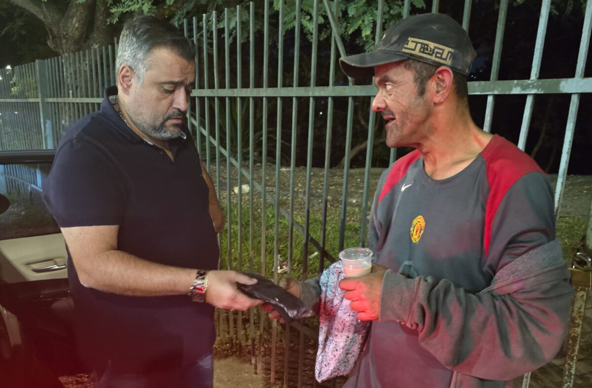 Dois homens estão interagindo em uma calçada à noite, próximo a uma grade metálica e árvores ao fundo. À esquerda, um homem de pele clara, barba grisalha e cabelo curto, veste uma camisa polo preta luvas ao outro homem. À direita, o segundo homem, que aparenta estar em situação de rua, tem a pele mais escura e o rosto sujo, usa um boné preto, uma blusa cinza com detalhes vermelhos .Ele segura um copo plástico transparente com líquido e um cobertor enrolado no braço. Ambos estão focados no momento da entrega, demonstrando um gesto de solidariedade.