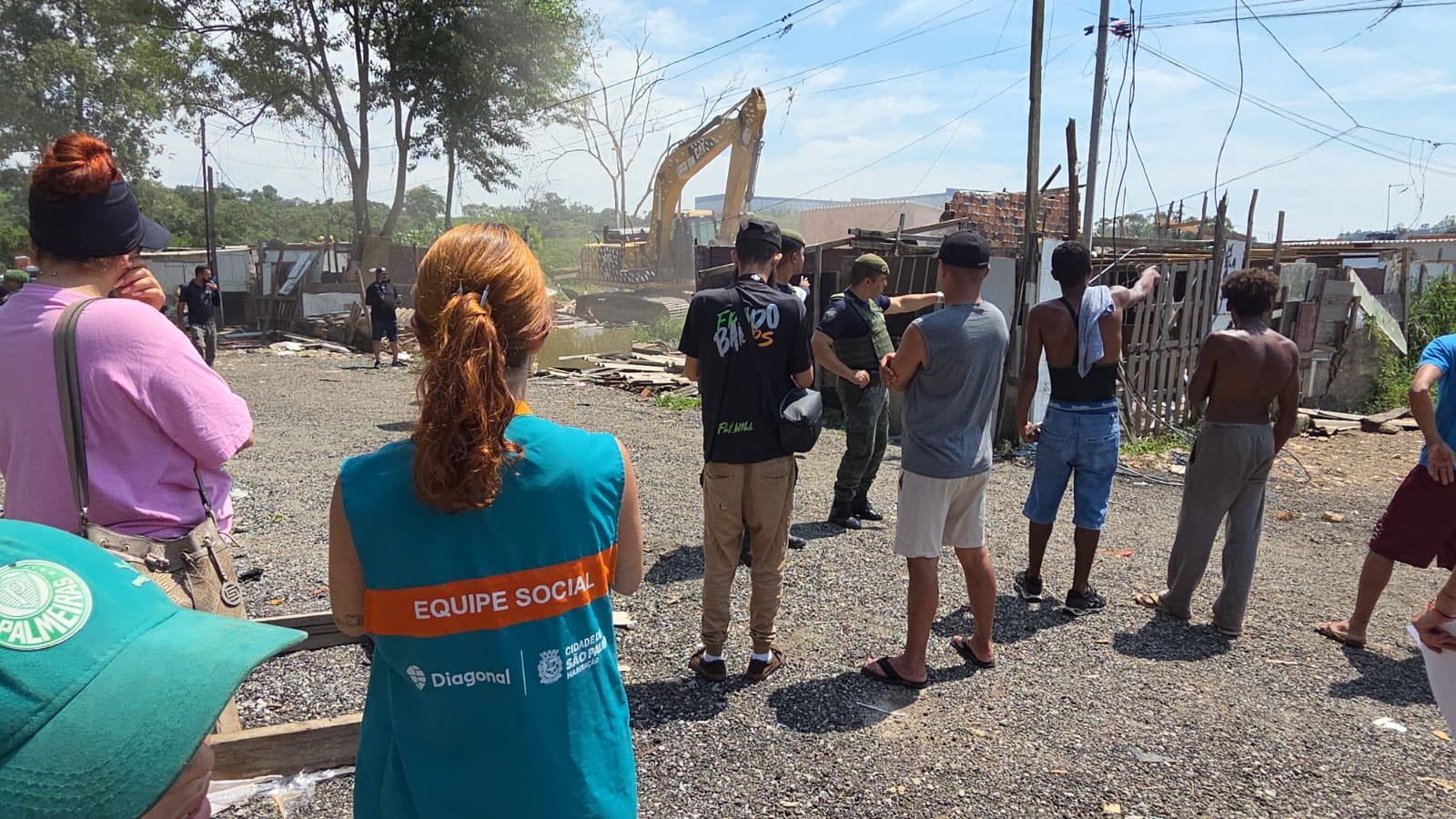 Pessoas de costas avistando a ação de trator realizando derrubada de barracos, dentre elas, à esquerda, uma mulher com colete azul e faixa laranja com a inscrição Equipe Social e logo da Secretaria Municipal de Habitação.