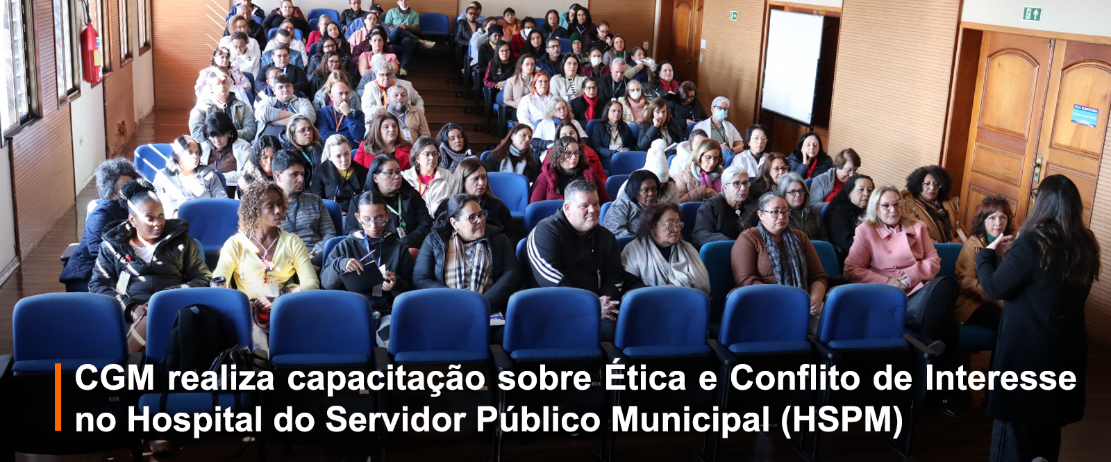 Sala pequena cheia de pessoas assistindo uma palestra