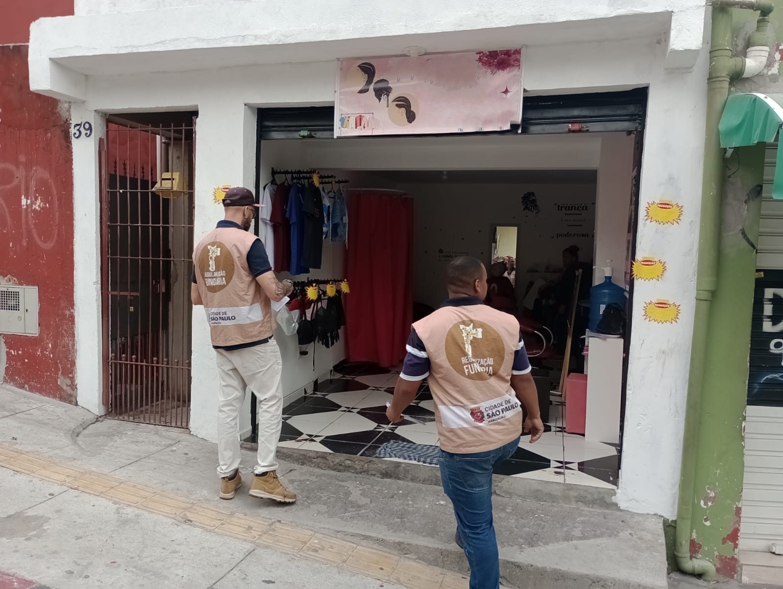 Dois homens de coletes bege da Regularização Fundiária – Cidade de São Paulo caminham pela calçada em direção à entrada de um pequeno comércio de roupas e salão de beleza. O espaço tem piso preto e branco em formato de losangos e uma cortina vermelha ao fundo. Na parede externa branca há adesivos amarelos em forma de sol e, ao lado esquerdo, o número 39 em azul. Um dos homens usa boné, máscara e botas claras; o outro veste calça jeans e tênis escuros.