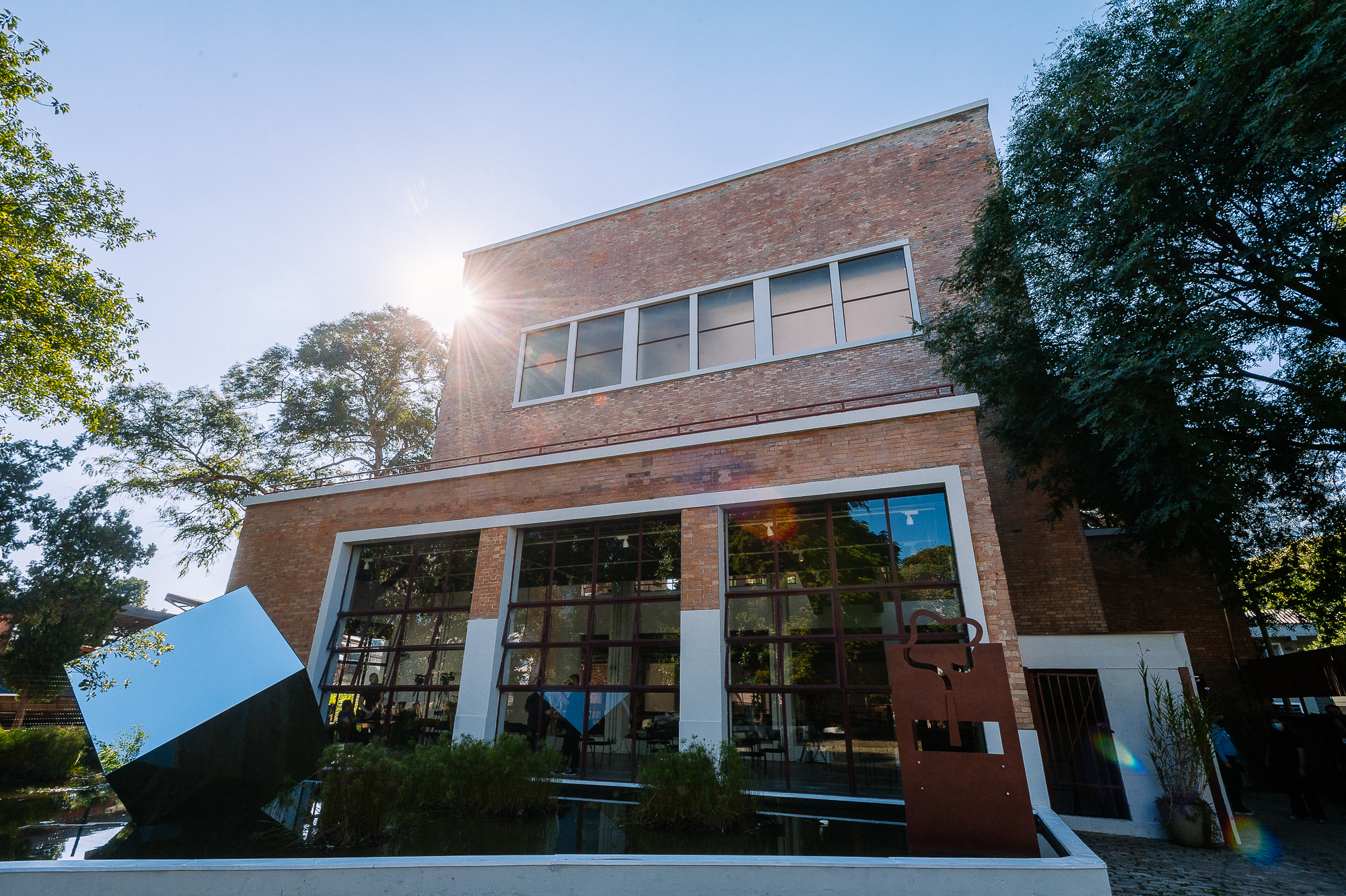 Fotografia colorida de um prédio de dois andares, feito de tijolos avermelhados e janelas grandes com molduras brancas. O sol aparece no canto superior esquerdo, brilhando por trás das árvores e criando um feixe de luz sobre a fachada. Em frente ao prédio há um espelho d’água com vegetação e duas esculturas geométricas: uma estrutura cúbica azul refletiva inclinada e uma peça marrom de metal recortado. Árvores verdes cercam o local sob um céu azul claro.