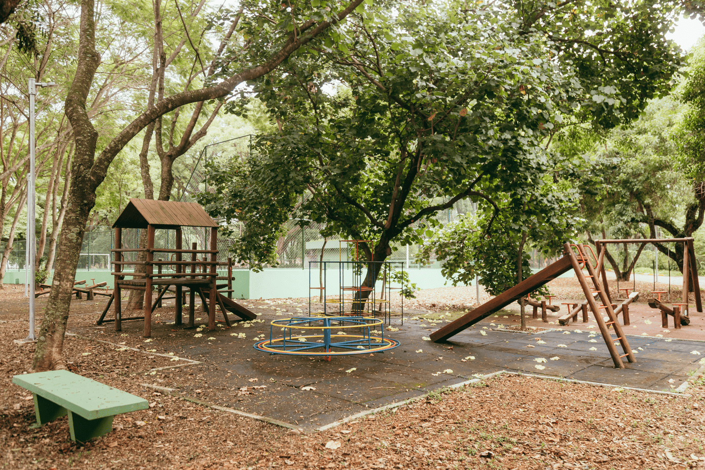 Parquinho infantil ao ar livre em área arborizada. Há vários brinquedos de madeira, como uma casinha elevada com escada e escorregador, além de um gira-gira colorido no centro. Ao lado, há outro escorregador e estruturas de equilíbrio. O chão é coberto por piso emborrachado e folhas secas, com árvores grandes ao redor criando sombra. Não há pessoas no local.