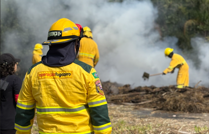 Em uma área de vegetação seca, brigadistas vestidos com uniformes amarelos e capacetes combatem um foco de incêndio em meio a muita fumaça branca. Em primeiro plano, um agente da Operação Fogo Zero observa a ação de costas, enquanto outros integrantes utilizam ferramentas para controlar as chamas e o material queimado no solo. Ao fundo, há árvores parcialmente encobertas pela fumaça.