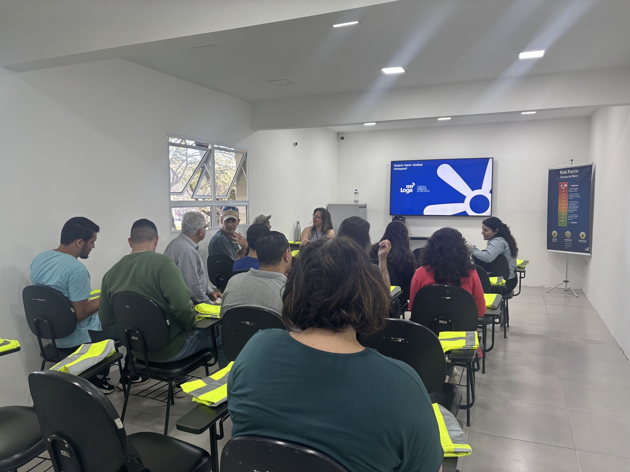 Participantes sentados em sala de treinamento interagem durante atividade, com apresentação projetada em tela sobre segurança.