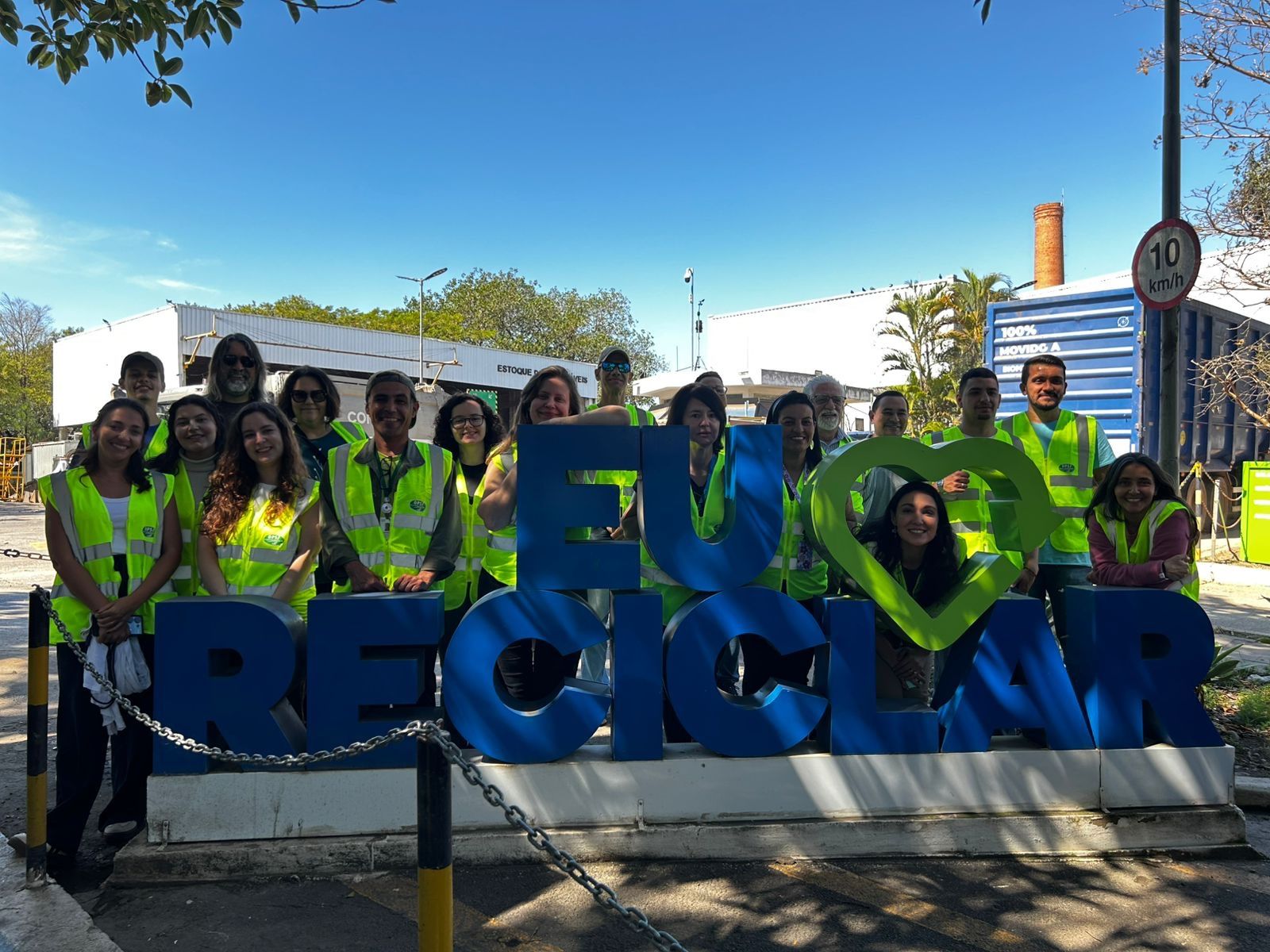 Grupo de pessoas usando coletes refletivos posam para foto em frente à escultura azul e verde com a frase “Eu ♥ Reciclar”.