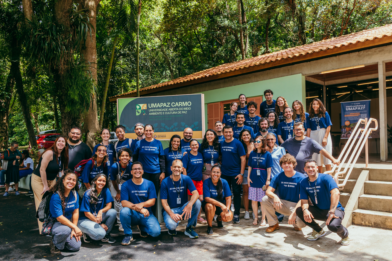Foto colorida ao ar livre, em área arborizada. Um grupo grande de cerca de 35 pessoas posa para a câmera em frente a um prédio claro, com telhado cerâmico, localizado em meio à mata. Todas vestem camisetas azuis com a inscrição “20 anos UMAPAZ”. As pessoas estão em pé e agachadas, sorrindo, organizadas em várias fileiras. Ao fundo, há uma placa verde com o texto “UMAPAZ Carmo – Universidade Aberta do Meio Ambiente e Cultura de Paz”. O ambiente é iluminado pela luz do dia, com árvores altas e vegetação densa ao redor, transmitindo clima de integração, celebração e contato com a natureza.