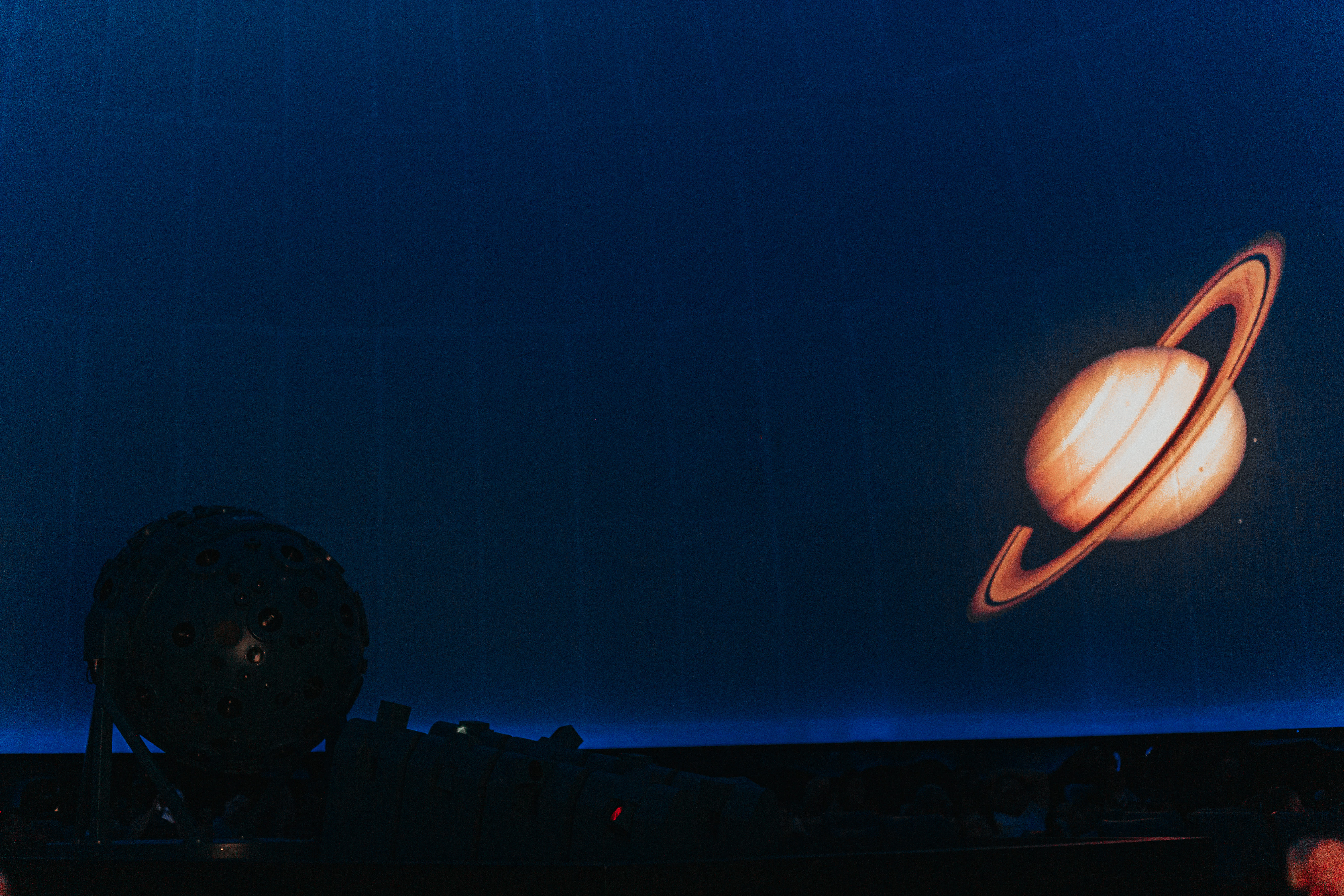 Interior de um planetário escuro. No centro da imagem, aparece a silhueta de um projetor esférico grande, cheio de pequenas lentes. Ele está iluminado apenas por reflexos suaves. Ao fundo, projetado na cúpula azul-escura, há a imagem de Saturno, com seus anéis alaranjados bem definidos. A cena transmite a sensação de imersão típica das sessões astronômicas, com o público sentado em silêncio no fundo, quase invisível na penumbra.