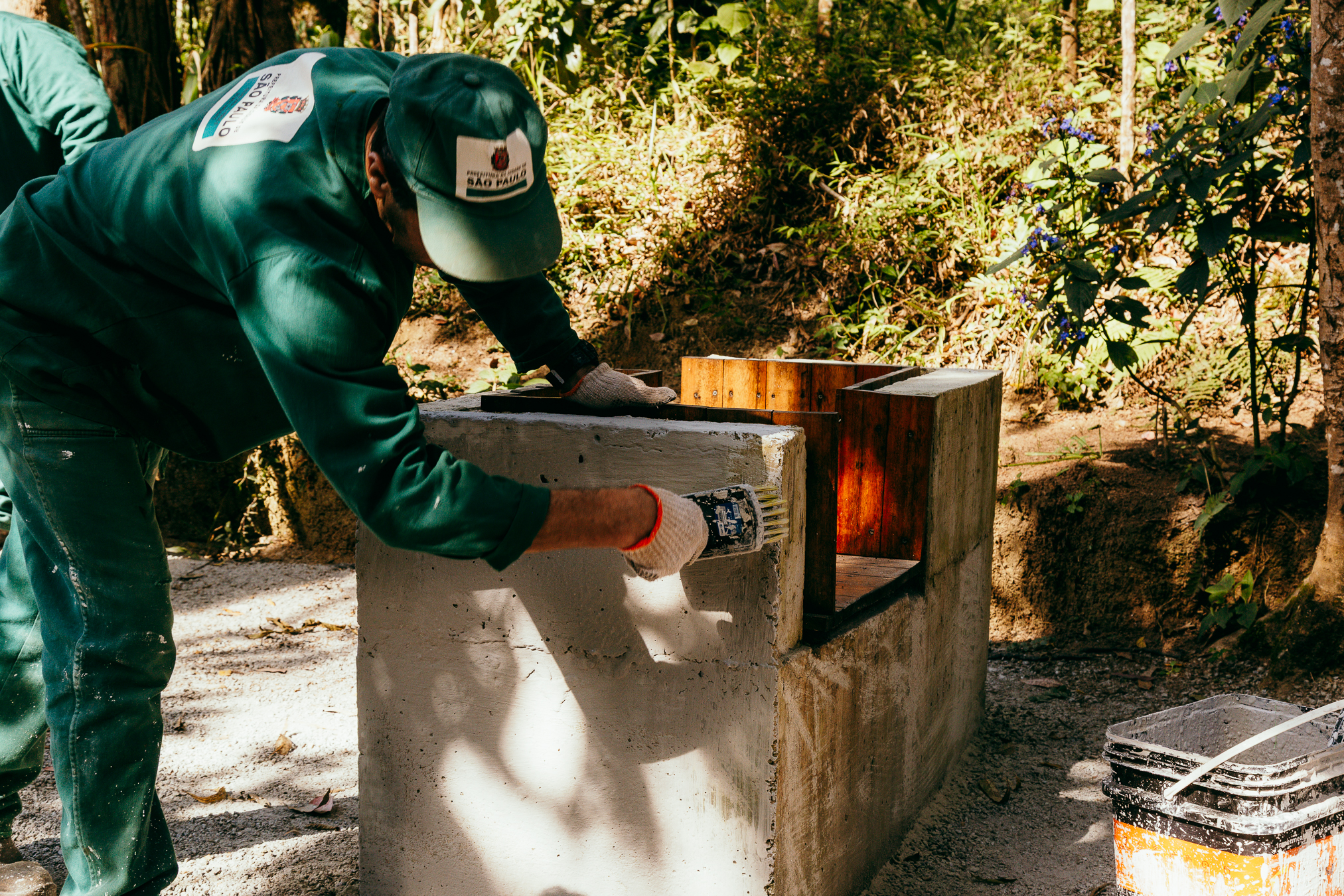 Trabalhador uniformizado com roupas verdes e boné do Governo do Estado de São Paulo aplica tinta ou impermeabilizante em uma estrutura de concreto recém-construída no meio da mata. Ao lado, há um balde com utensílios de pintura e, ao fundo, vegetação nativa.