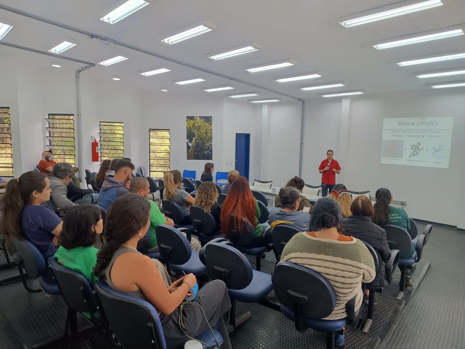 Palestrante apresenta conteúdo sobre riscos em uma sala de treinamento, enquanto diversas pessoas assistem atentas às explicações.