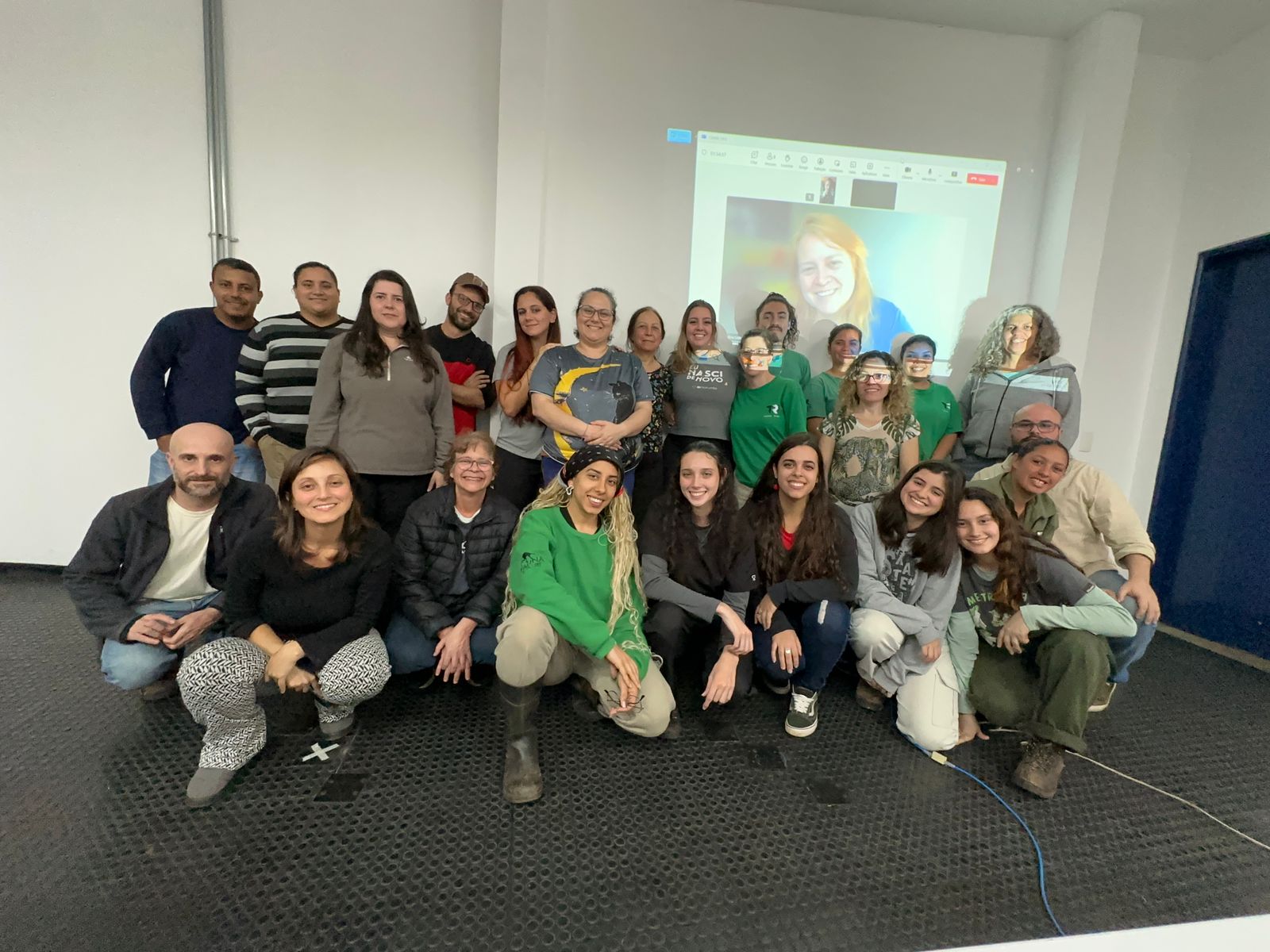 Grupo de participantes reunidos para foto coletiva em sala de treinamento, com projeção de uma participante em chamada online ao fundo.
