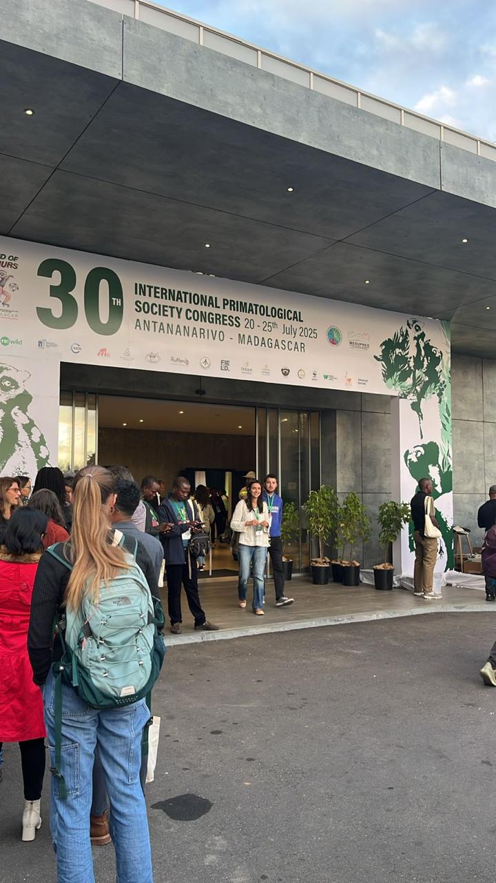 Uma fotografia de uma multidão de pessoas do lado de fora de um evento. Elas estão em frente à entrada de um prédio, que tem um grande banner verde e branco com a inscrição "30th International Primatological Society Congress" e "20-25th July 2025 Antananarivo - Madagascar". Há pessoas conversando e entrando no evento.