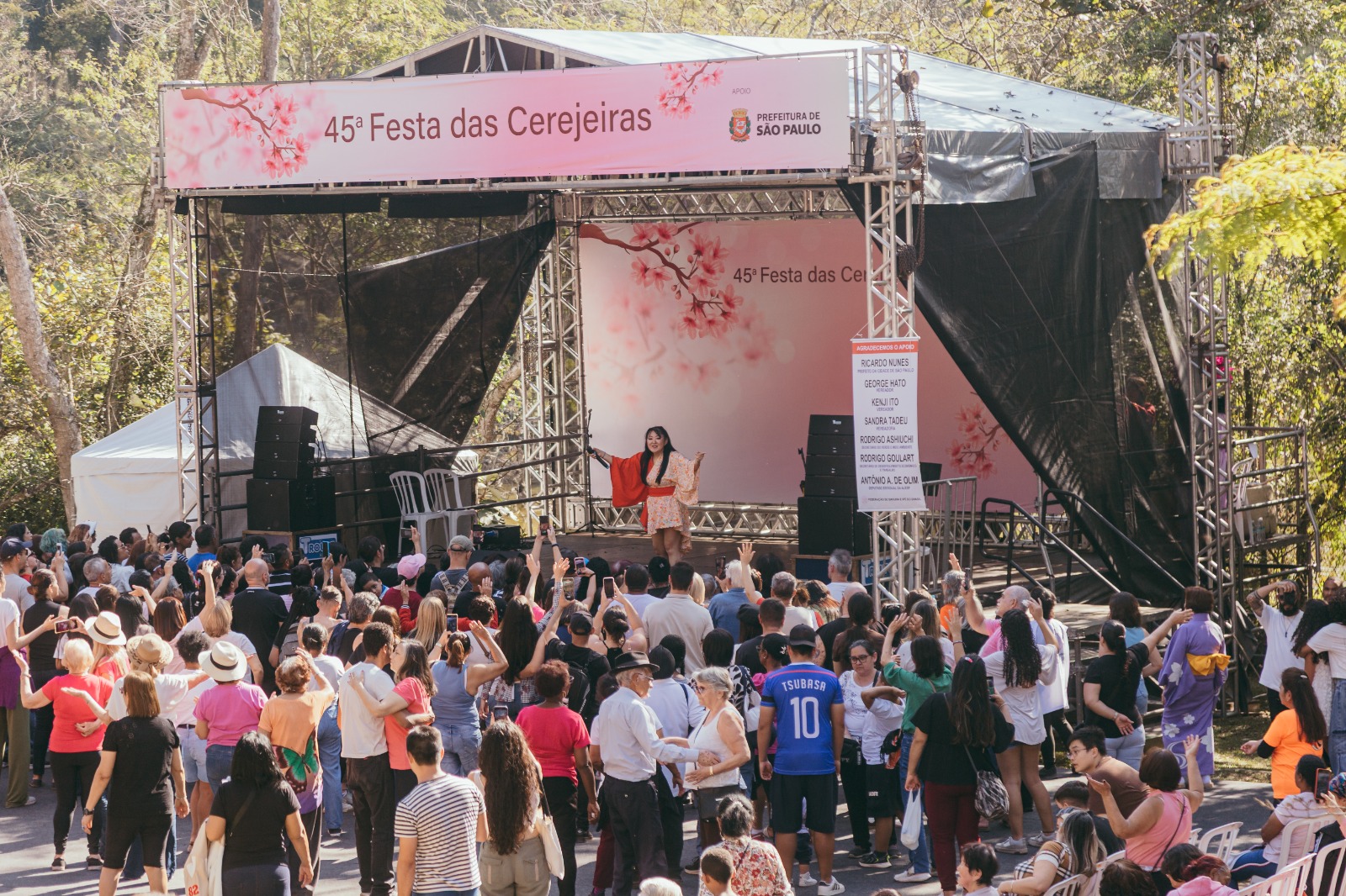 Multidão se reúne em frente a um palco montado para a 45ª Festa das Cerejeiras, promovida pela Prefeitura de São Paulo. Uma mulher com traje tradicional japonês se apresenta no palco decorado com imagem de flores de cerejeira. A plateia acompanha atenta e animada, registrando o momento com celulares. O ambiente é cercado por árvores, compondo um cenário festivo ao ar livre, típico de celebrações culturais e sazonais.