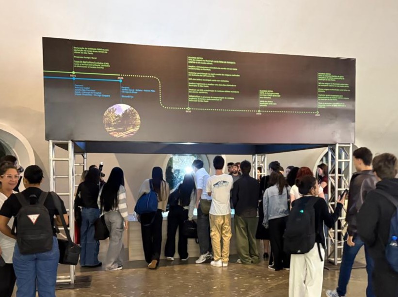 A imagem mostra um grupo diverso de pessoas em frente a uma estrutura expositiva com linha do tempo e área interativa, em um ambiente educativo e acessível.