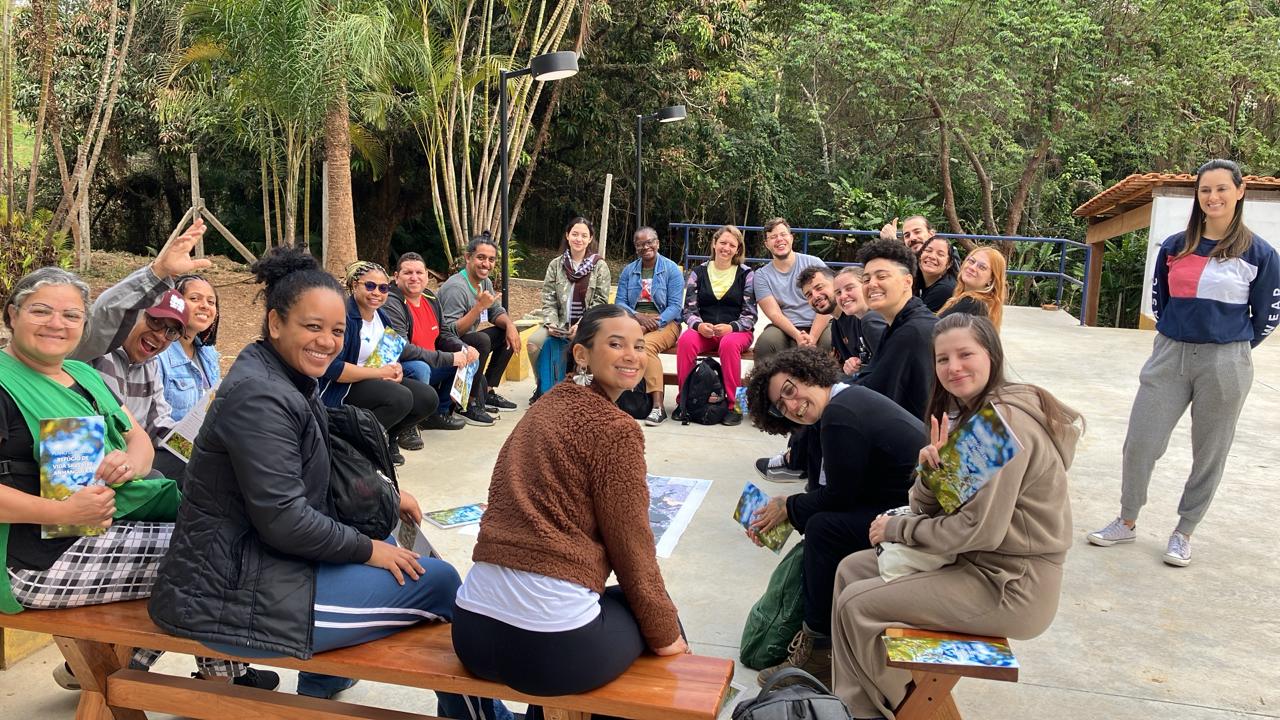 Grupo diverso de pessoas reunido ao ar livre, sentado em semicírculo, sorrindo e interagindo. Há árvores ao fundo e todos parecem participar de uma atividade com livros ou cadernos nas mãos. Ambiente acolhedor e natural.