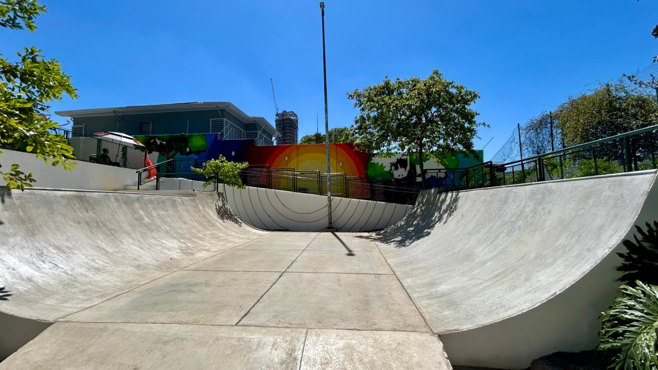 A imagem mostra uma pista de skate em formato de “U”, feita de concreto liso e cinza-claro, sob um céu azul intenso e ensolarado. Ao fundo, há um muro colorido com um grande arco-íris pintado, que vai do vermelho ao violeta. Nas laterais do muro, aparecem desenhos de árvores e animais em tons de verde, preto e branco. À esquerda, há uma pequena sombra de um guarda-sol branco e vermelho. Árvores com folhagem verde clara e escura completam a paisagem, trazendo contraste com o cinza da pista e o azul vibrante do céu.