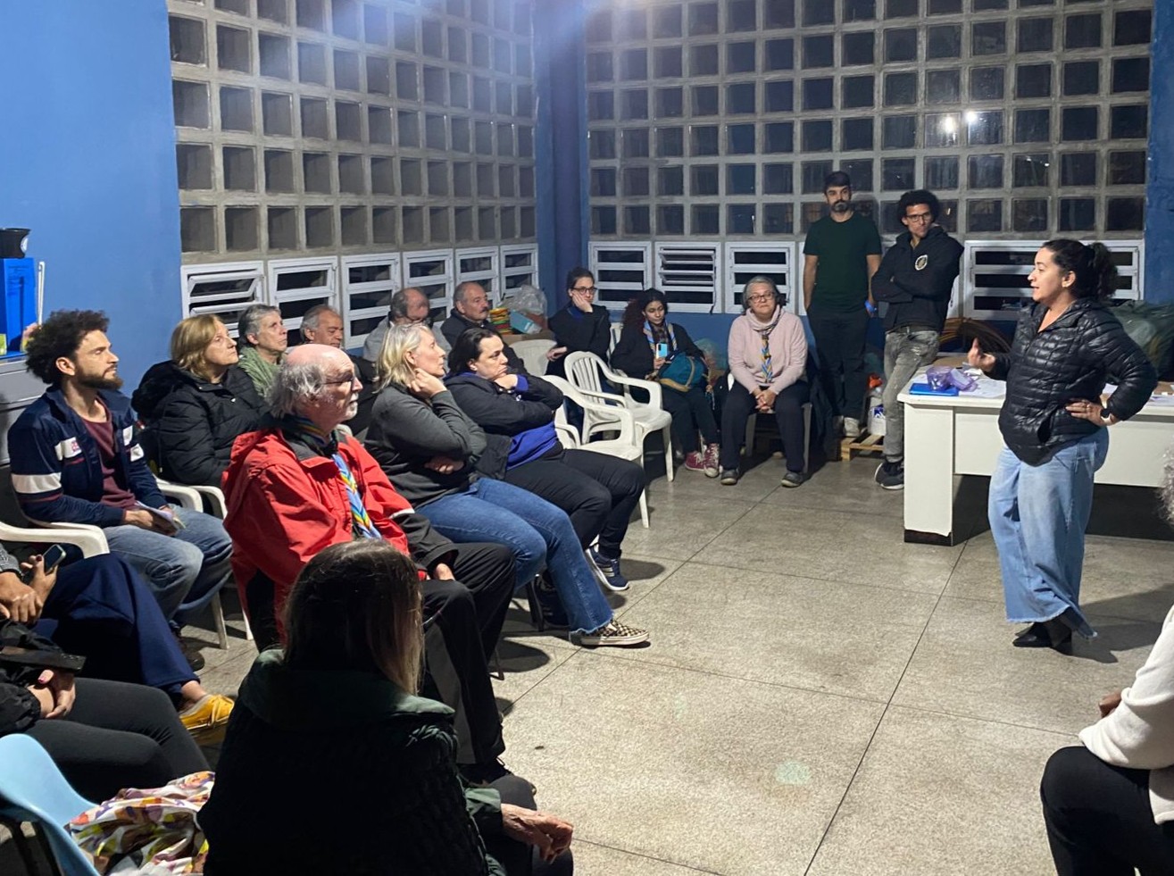 Pessoas participam de uma reunião em uma sala com paredes azuis. Um grupo está sentado em cadeiras plásticas, atento a uma mulher em pé que fala na frente do espaço. O ambiente é iluminado por lâmpadas pendentes e tem janelas de vidro blocado.