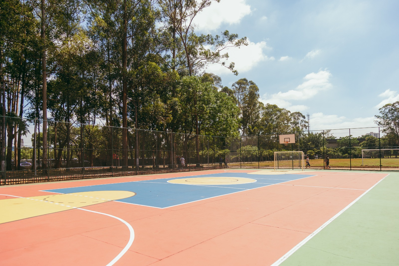 Quadra poliesportiva ao ar livre, com piso colorido em tons de laranja, azul, verde e amarelo, demarcado para diferentes modalidades. Há traves de futebol e cestas de basquete nas extremidades. A quadra é cercada por alambrado e está localizada em uma área arborizada, com muitas árvores altas ao redor. Ao fundo, vê-se um campo gramado e algumas pessoas caminhando. O céu está claro, com poucas nuvens, indicando um dia ensolarado.