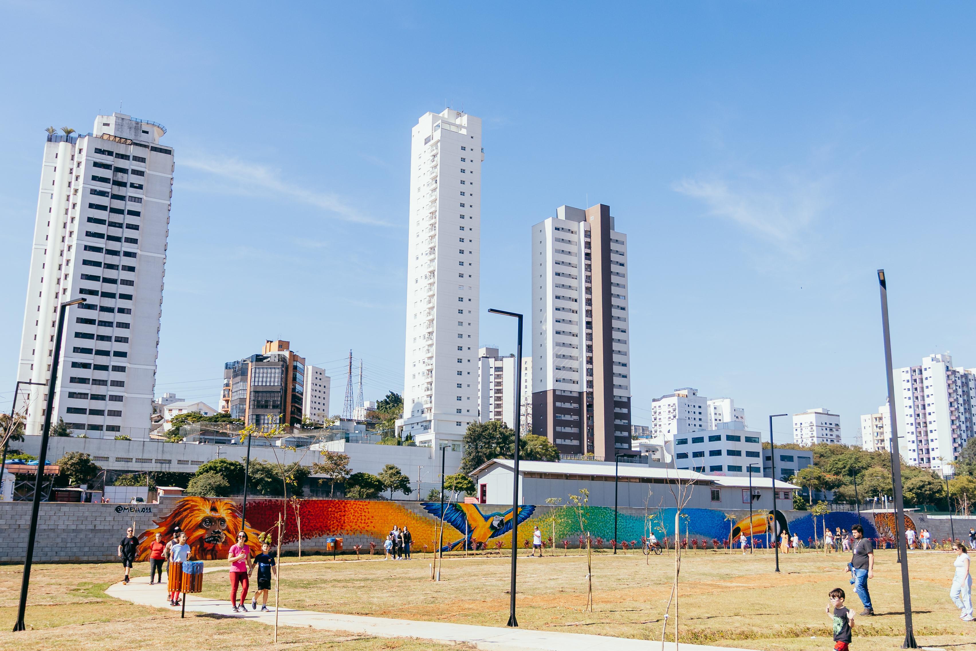 A imagem mostra uma ampla área aberta de parque urbano em um dia ensolarado, sob um céu azul-claro praticamente sem nuvens. Em primeiro plano, há gramados em tons de verde e amarelo-palha, cortados por uma calçada sinuosa de concreto claro. Algumas pessoas caminham pelo parque, incluindo adultos e crianças, enquanto outras passeiam ou observam o espaço. Postes de iluminação pretos e árvores jovens recém-plantadas estão distribuídos pelo terreno, indicando uma área de uso público em processo de consolidação paisagística.  Ao fundo, um longo muro é coberto por um mural artístico colorido que retrata animais da fauna brasileira. Destacam-se um mico-leão-dourado em tons vibrantes de laranja e dourado, uma arara de penas azuis e amarelas com as asas abertas e um tucano com bico amarelo e preto. O mural apresenta um degradê de cores intensas, passando pelo vermelho, laranja, amarelo, verde e azul. Atrás do muro, ergue-se o cenário urbano composto por edifícios residenciais altos, predominantemente brancos e cinzas, contrastando com a área verde do parque. A iluminação natural intensa valoriza as cores vivas do mural e destaca a paisagem que integra natureza, arte urbana e verticalização da cidade.