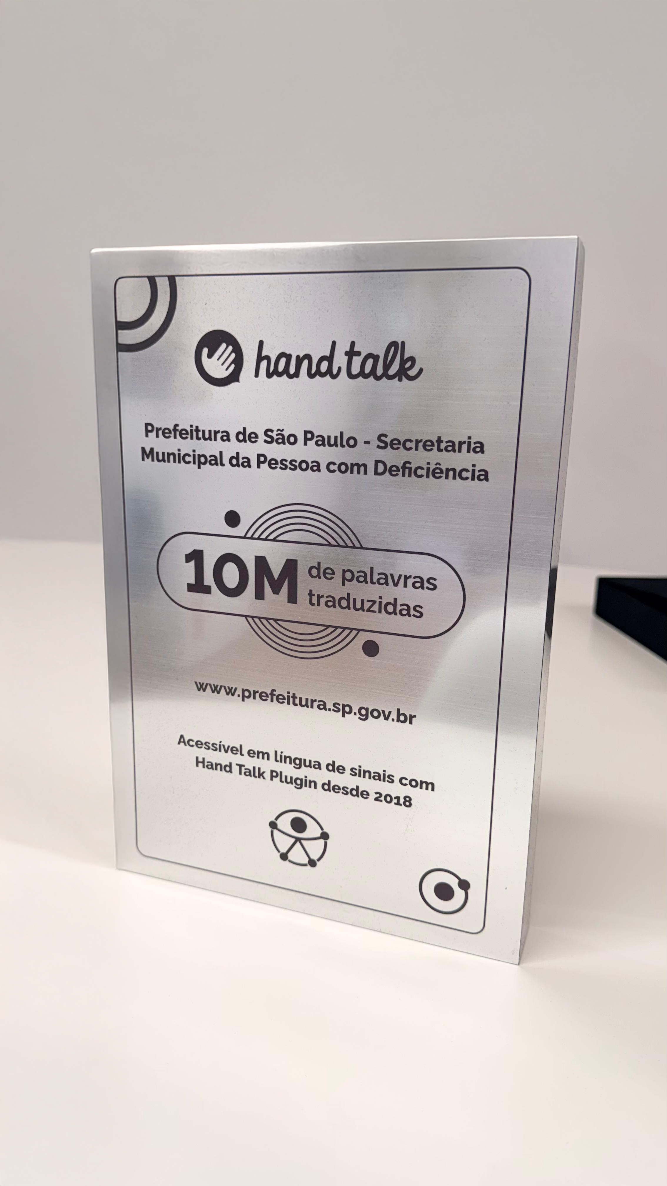 Placa de aço inox. No topo, logotipo do Hand Talk. Abaixo, o seguinte texto: Prefeitura de São Paulo. Secretaria Municipal da Pessoa com Deficiência. 10 milhões de palavras traduzidas. www.prefeitura.sp.gov.br. Acessível em língua de sinais com Hand Talk Plugin desde 2018. No final, o símbolo de acessibilidade universal. 
