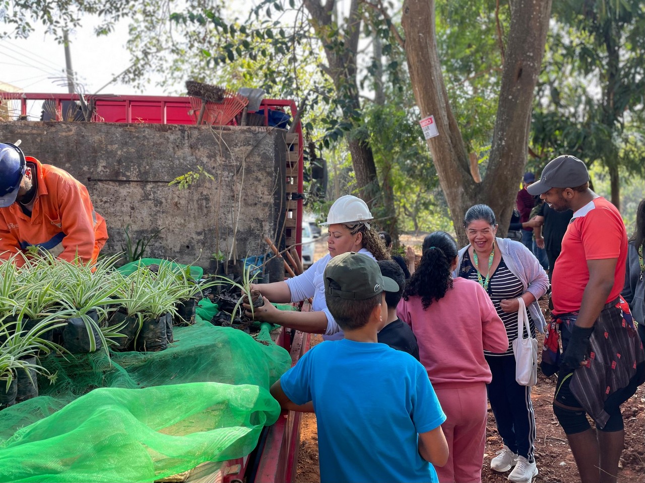 #PraTodosVerem: Caminhão carregando mudas de plantas, homem de uniforme laranja trabalhando. Ao lado, pessoas conversando e mulher de branco com capacete mexendo nas plantas do caminhão.