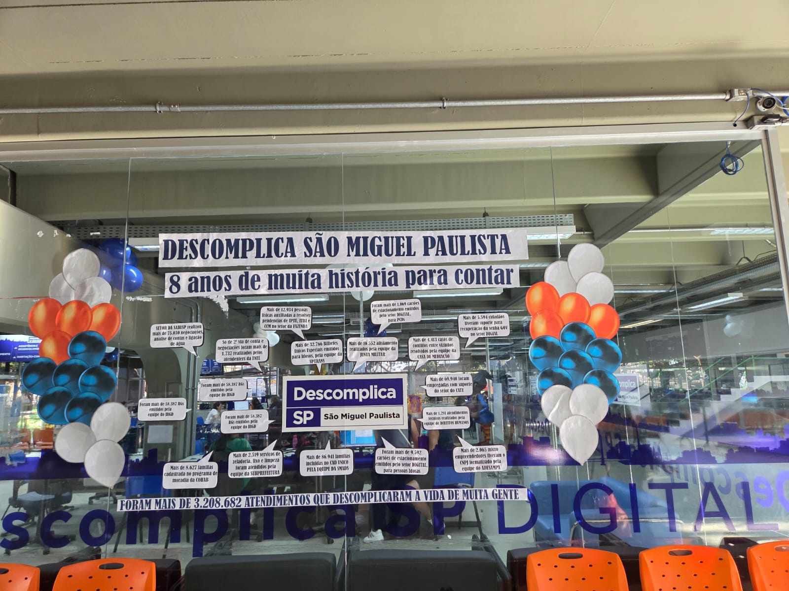 Fachada de vidro do Descomplica São Miguel Paulista decorada com balões e adesivos comemorativos de 8 anos, destacando a marca de mais de 3,2 milhões de atendimentos realizados.
