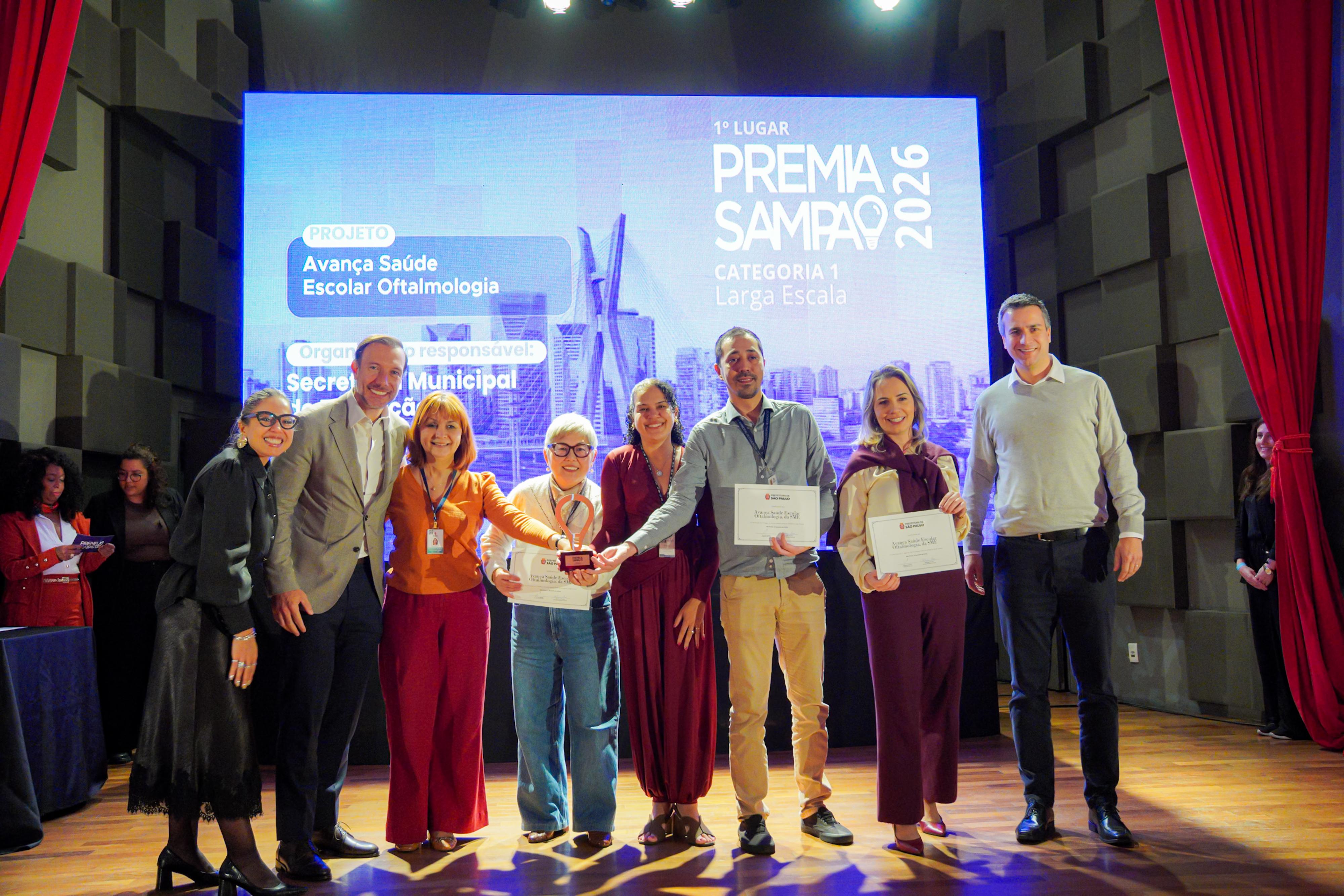 A fotografia registra uma cerimônia de premiação realizada em um auditório, com oito representantes da Secretaria Municipal da Saúde de São Paulo posando no palco após receberem um reconhecimento institucional. Ao centro, integrantes da equipe exibem certificados e um troféu, enquanto os demais participantes celebram a conquista.  No grande telão ao fundo, é possível identificar que o projeto premiado foi o “Avança Saúde Escolar Oftalmologia”, vencedor do 1º lugar no Premia Sampa 2026, na Categoria 1 – Larga Escala. A premiação reconhece iniciativas de destaque desenvolvidas pela administração municipal.  Os participantes aparecem sorridentes e em clima de comemoração, reunidos diante do painel da premiação. O palco é emoldurado por cortinas vermelhas e iluminado por refletores, reforçando o caráter solene e festivo do evento.