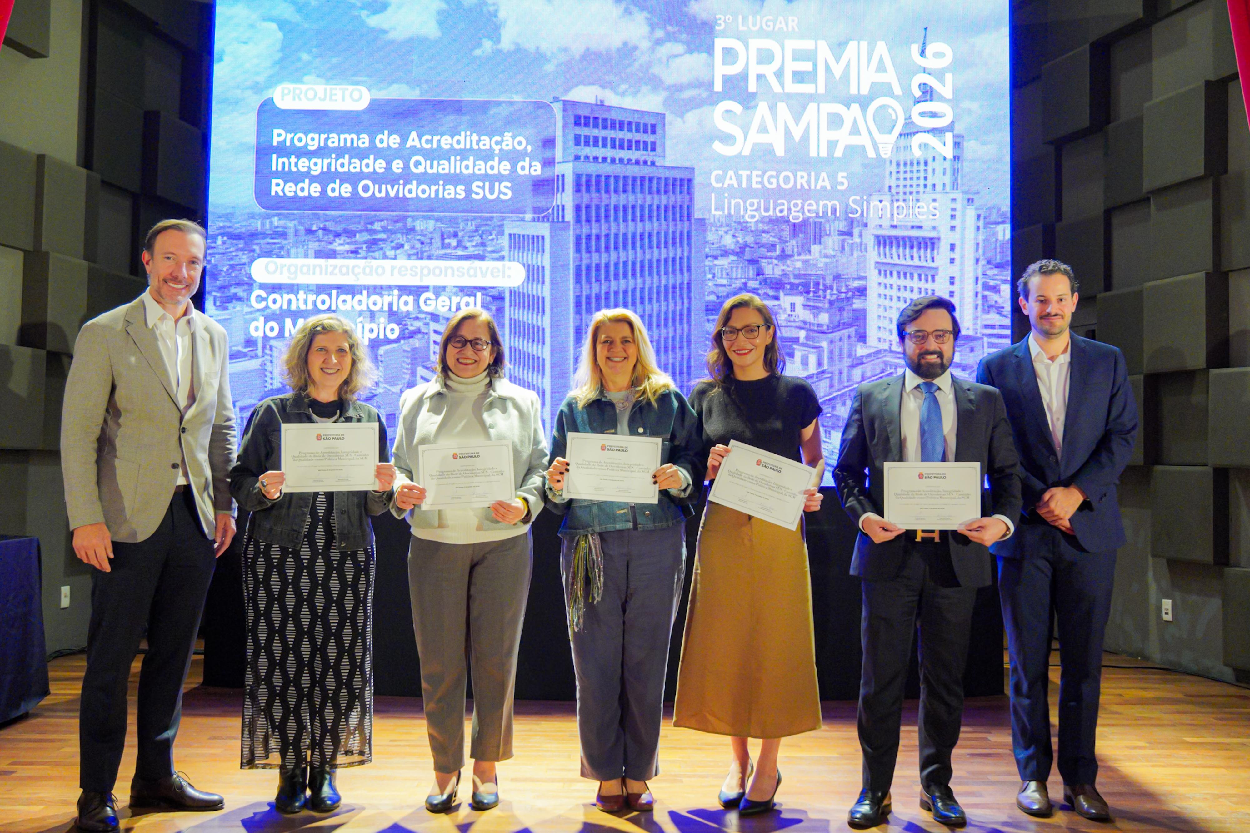 A fotografia mostra uma cerimônia de premiação realizada em um auditório, com sete participantes posando no palco diante de um grande telão que exibe os resultados do Premia Sampa 2026. Os integrantes seguram certificados de reconhecimento e aparecem sorridentes, em um momento de celebração institucional.  Ao fundo, o telão informa que o projeto premiado foi o “Programa de Acreditação, Integridade e Qualidade da Rede de Ouvidorias SUS”, contemplado com o 3º lugar na Categoria 5 – Linguagem Simples. Também é possível identificar a Controladoria Geral do Município como órgão responsável pela iniciativa.  Os participantes estão alinhados no centro do palco, vestindo trajes sociais e casuais, enquanto dois representantes da organização aparecem nas extremidades da foto. A iluminação cênica e o painel digital reforçam o caráter formal da cerimônia de reconhecimento de boas práticas da administração pública municipal.