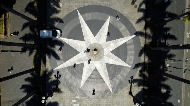 Vista aérea vertical de uma praça pública com um grande desenho de estrela de 8 pontas em tons de cinza e branco no pavimento.  O desenho central, que se assemelha a uma rosa dos ventos estilizada, domina a imagem. O centro da estrela é marcado por uma estrutura elevada e octogonal, onde algumas pessoas estão em pé.  Em torno da estrela, o chão é escuro. Há palmeiras altas em ambos os lados, e suas longas sombras escuras se projetam radialmente sobre a praça.  Diversas pessoas, cujas sombras também são visíveis, caminham pelo local. Um carro branco e preto (possivelmente uma viatura policial) está estacionado no canto superior esquerdo do desenho. Há postes de luz em estilo antigo e moderno ao redor da praça.  A iluminação é forte, sugerindo um dia de sol.