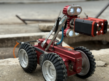 Fotografia em close-up de um robô de inspeção técnica sobre uma superfície de concreto. O dispositivo possui um corpo robusto em metal vermelho e quatro rodas grandes com pneus de borracha pretos e sulcos profundos para tração. Na parte superior, um braço mecânico articulado sustenta uma cabeça móvel equipada com uma câmera central e quatro potentes faróis de LED circulares (dois acima e dois abaixo da lente) que estão acesos, emitindo uma luz branca brilhante. Um cabo laranja está conectado à parte traseira do robô. O fundo da imagem está desfocado, mostrando de forma sutil elementos de uma calçada e uma via urbana.
